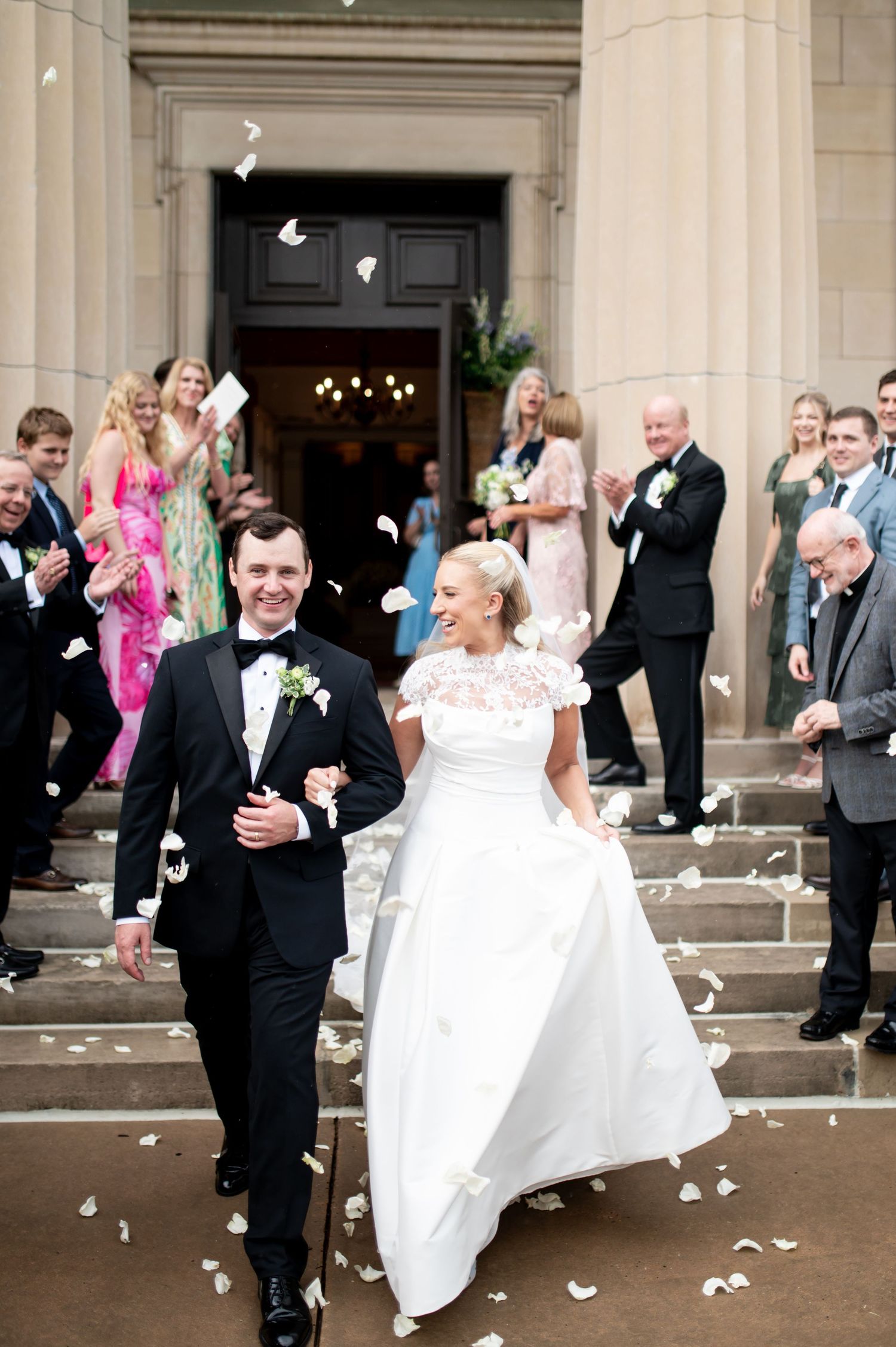 Bride and groom exit church ceremony on steps as guests toss white rose petals in celebration.