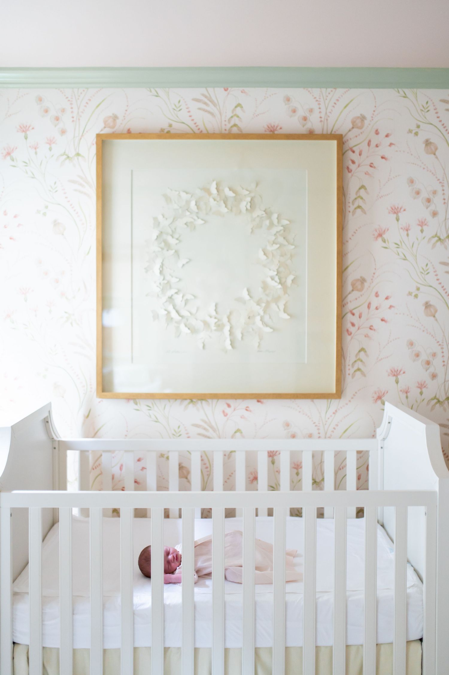 A white nursery crib against a wall with delicate pink floral wallpaper and framed artwork above.