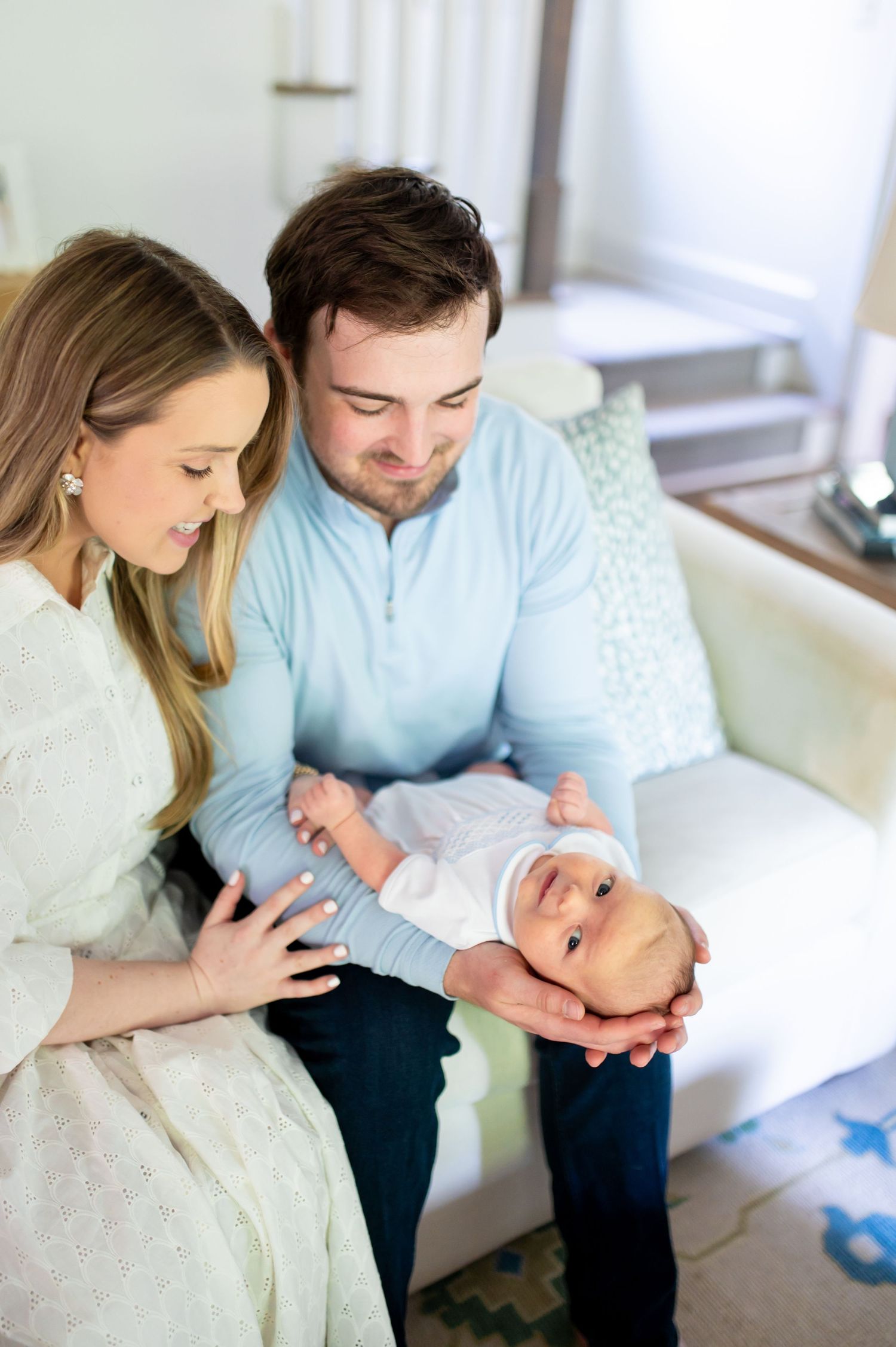 A couple sits on a white sofa, lovingly gazing at their newborn baby cradled in the father's hands.