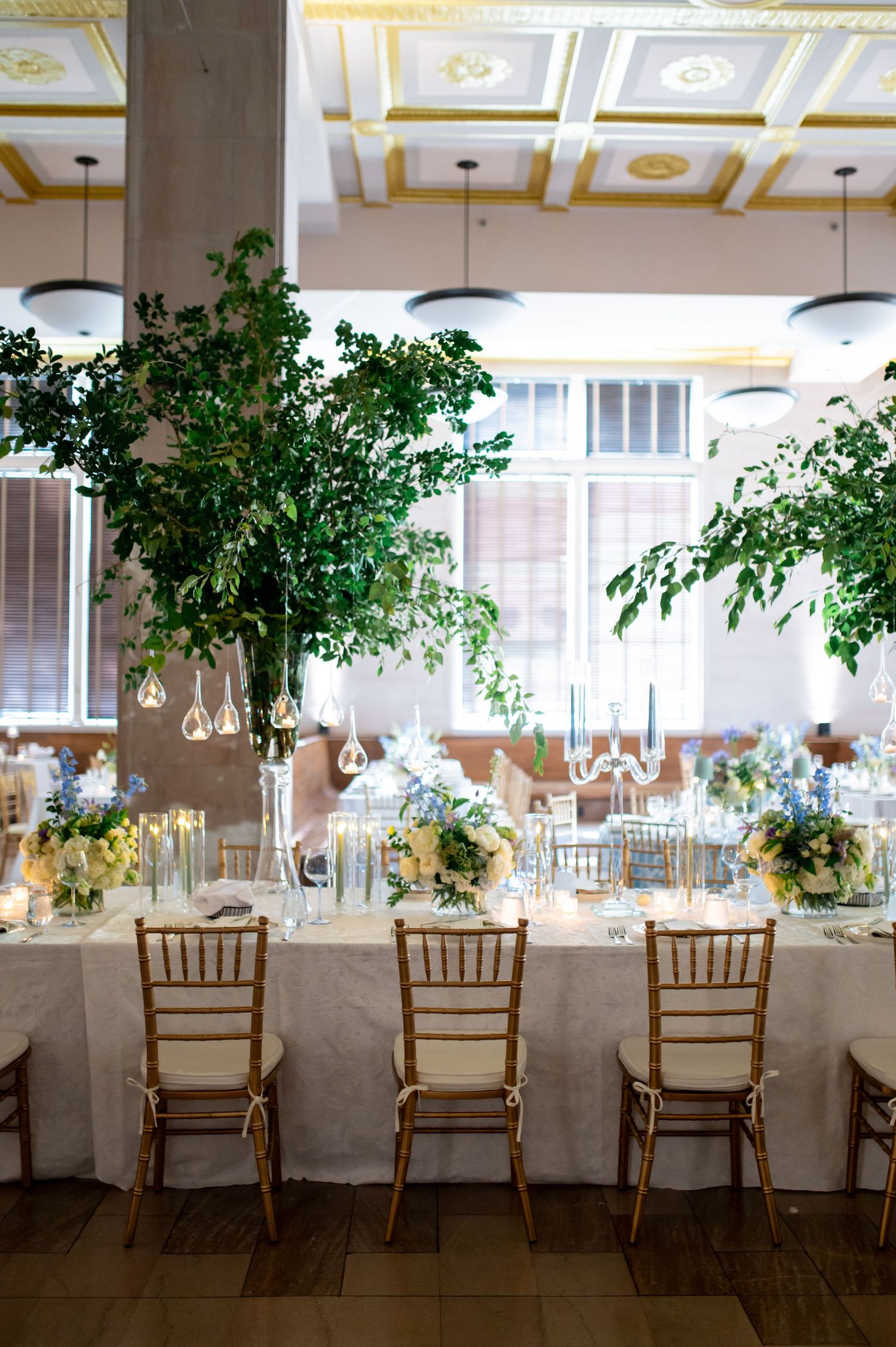 Elegant wedding reception with gold Chiavari chairs, white linen tables, lush greenery centerpieces, and soft floral arrangements.