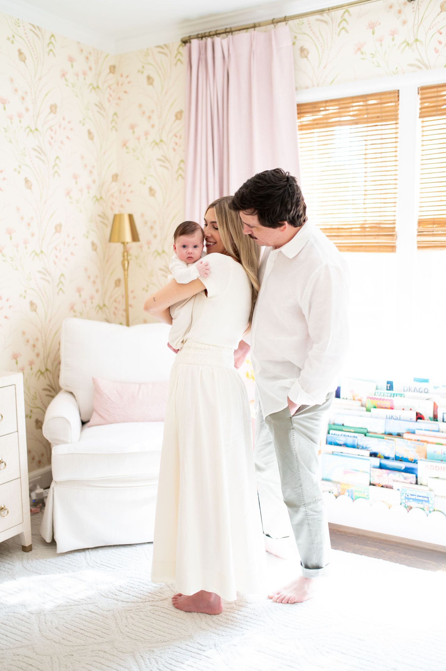 A couple in white clothing shares a tender moment while holding their newborn baby in a bright, floral nursery.