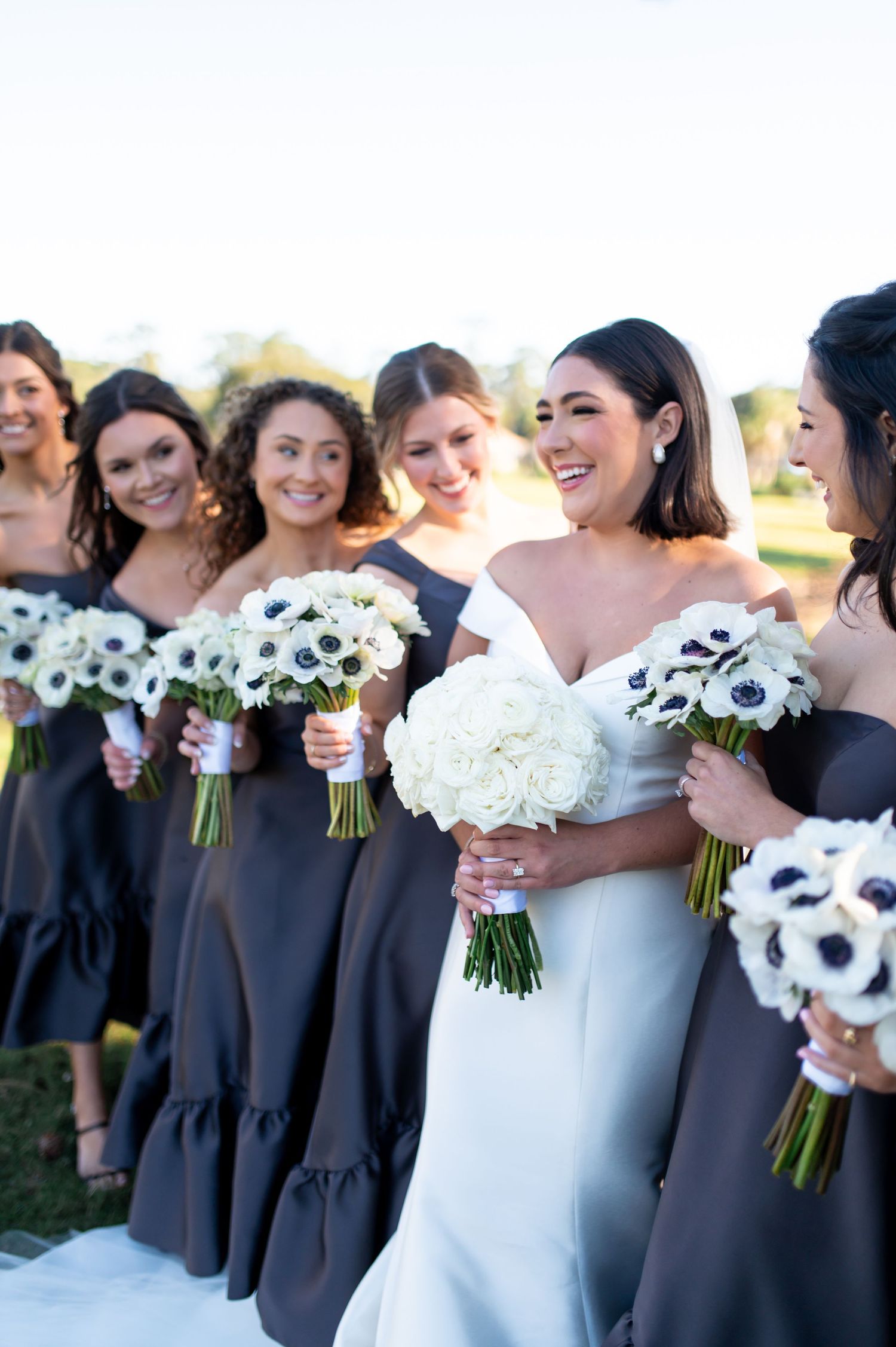 Bridesmaids in navy dresses hold black and white anemone bouquets at an outdoor wedding.
