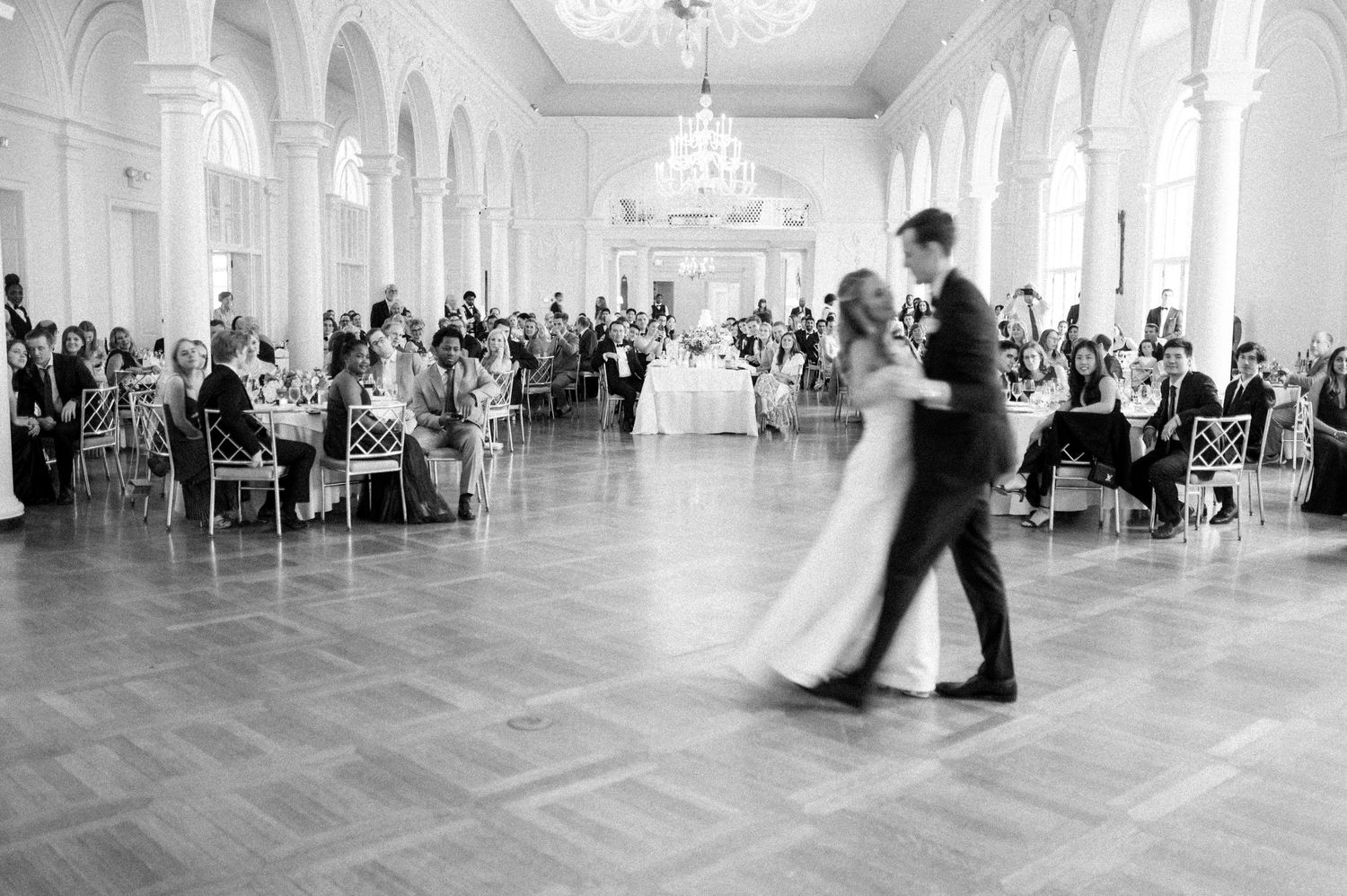 Black and white panorama of wedding guests watching first dance in grand ballroom.