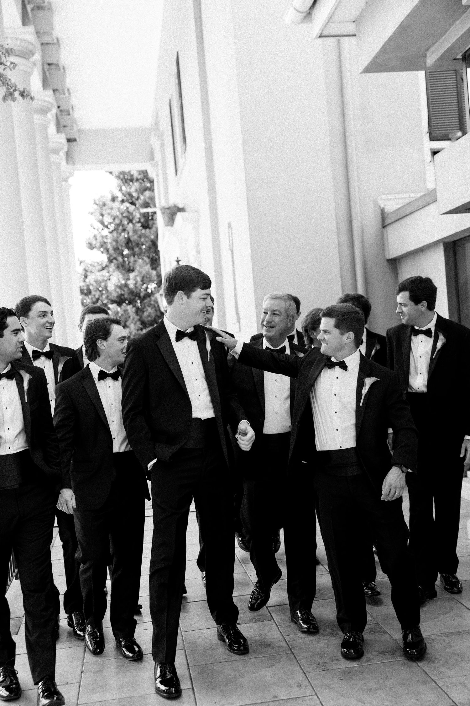 Groomsmen in black tuxedos laughing and walking together outdoors in a black and white wedding photo.