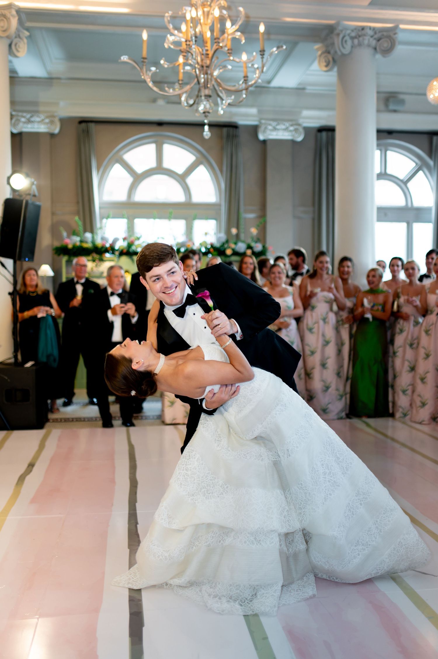 Groom dips bride dramatically on dance floor during first dance while wedding guests cheer in elegant ballroom.