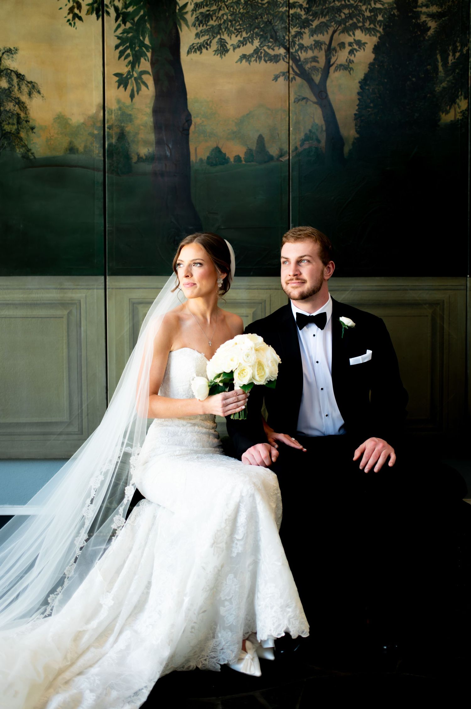 An elegant wedding couple shares an intimate moment on a settee with a scenic mural background.