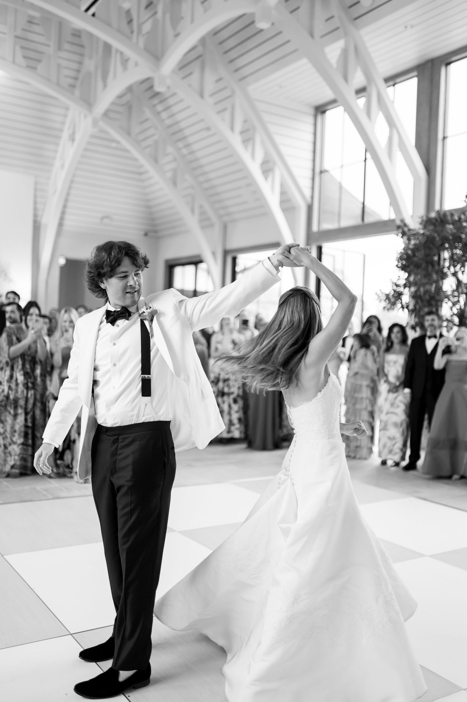 Black and white photo of newlyweds dancing in a grand ballroom surrounded by wedding guests.