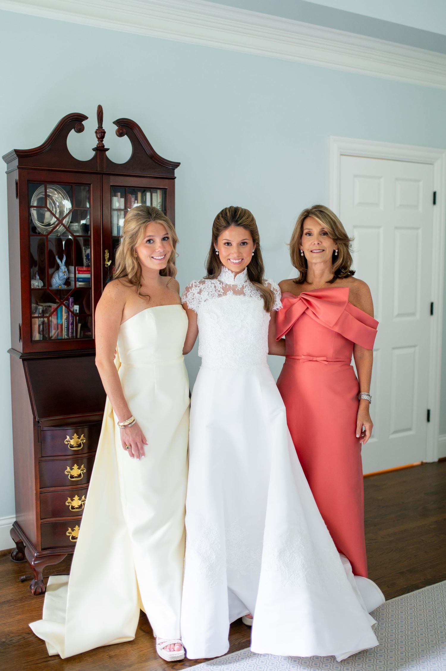 Three women pose with wedding dresses in an elegant room with antique furniture.