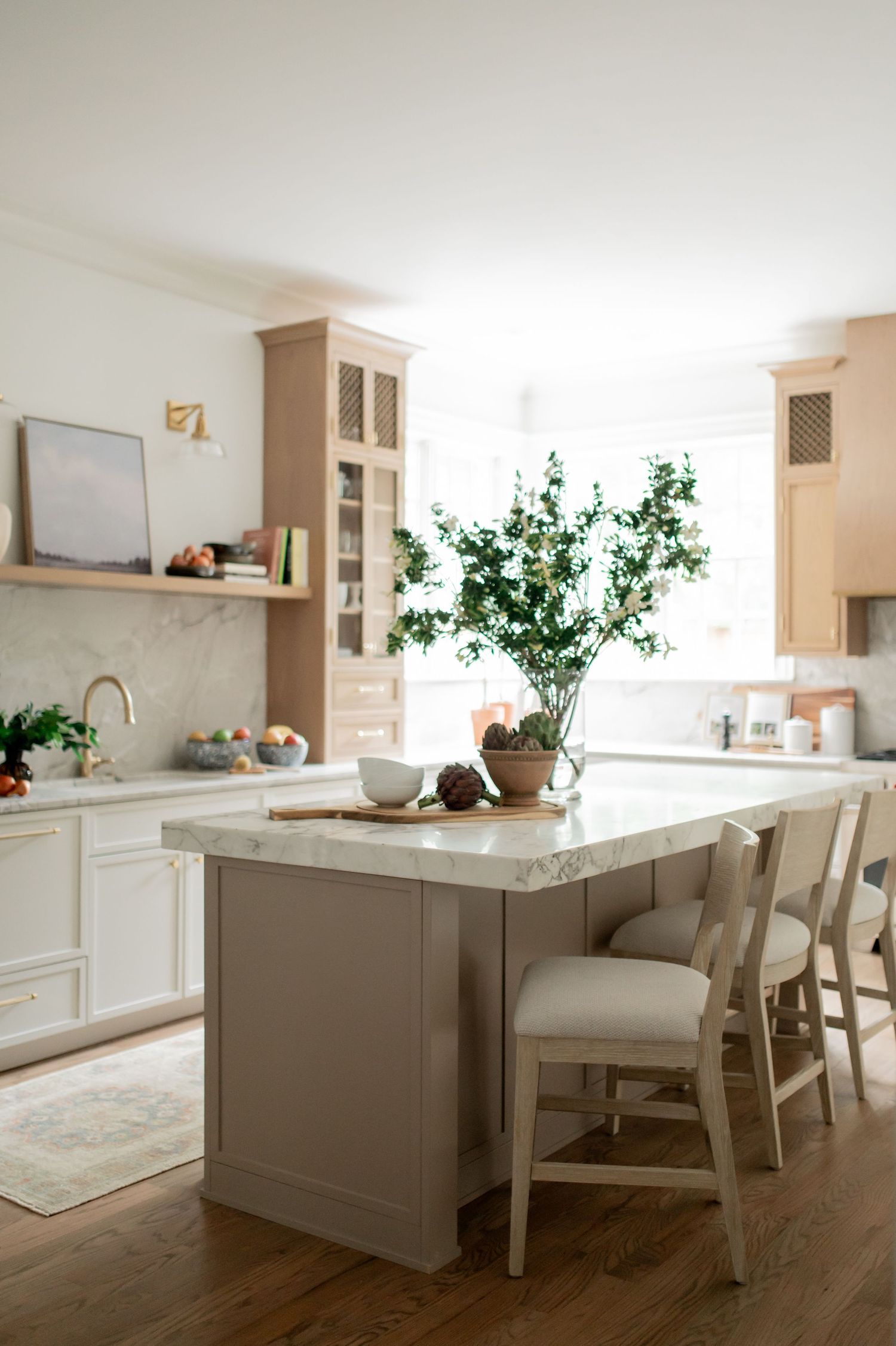 Bright kitchen with taupe island, marble countertops, white cabinets, light wood accents, and a green branch centerpiece.
