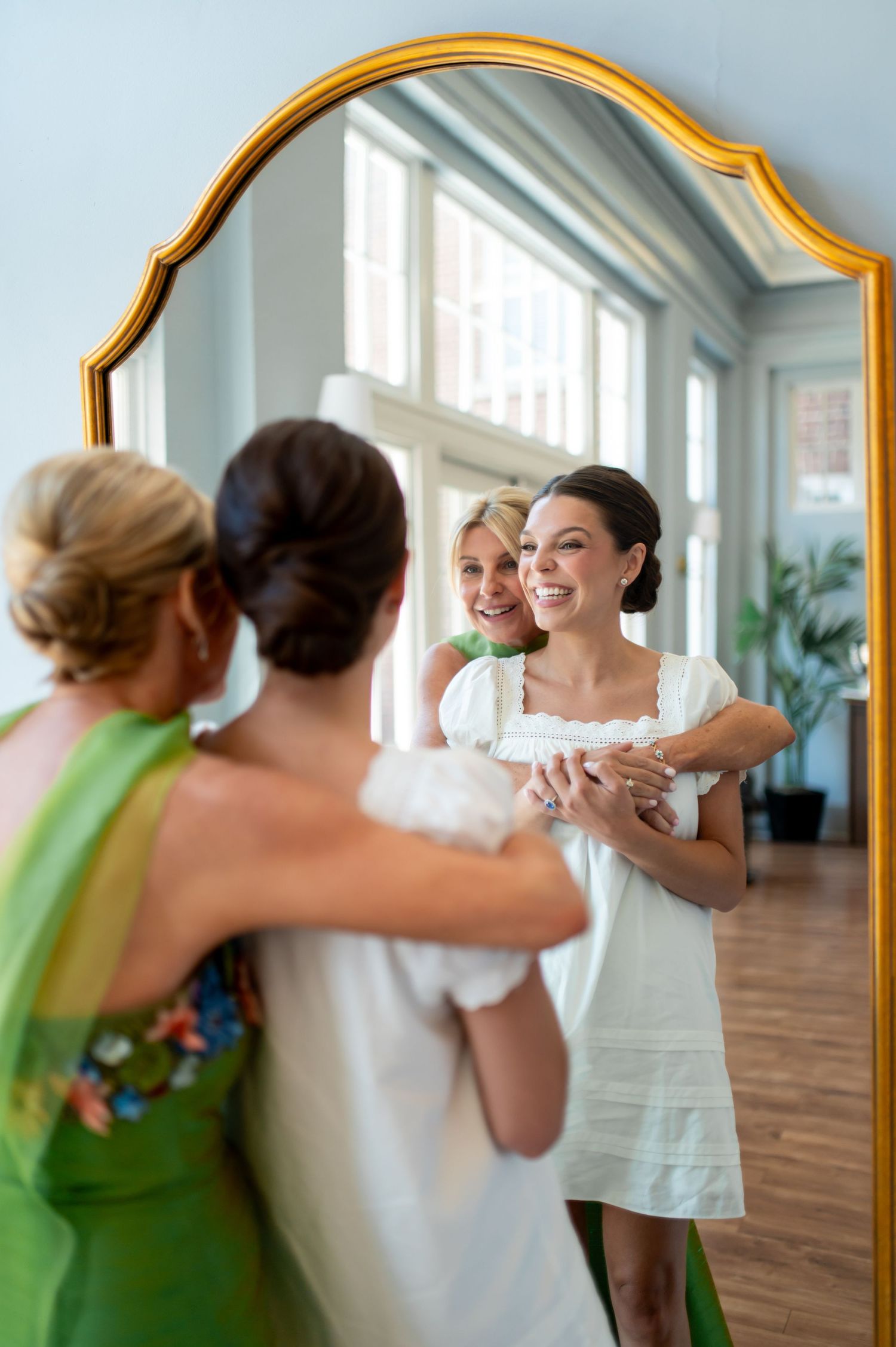 Bride in white dress smiles in gold mirror reflection with bridesmaids in green during wedding preparations.