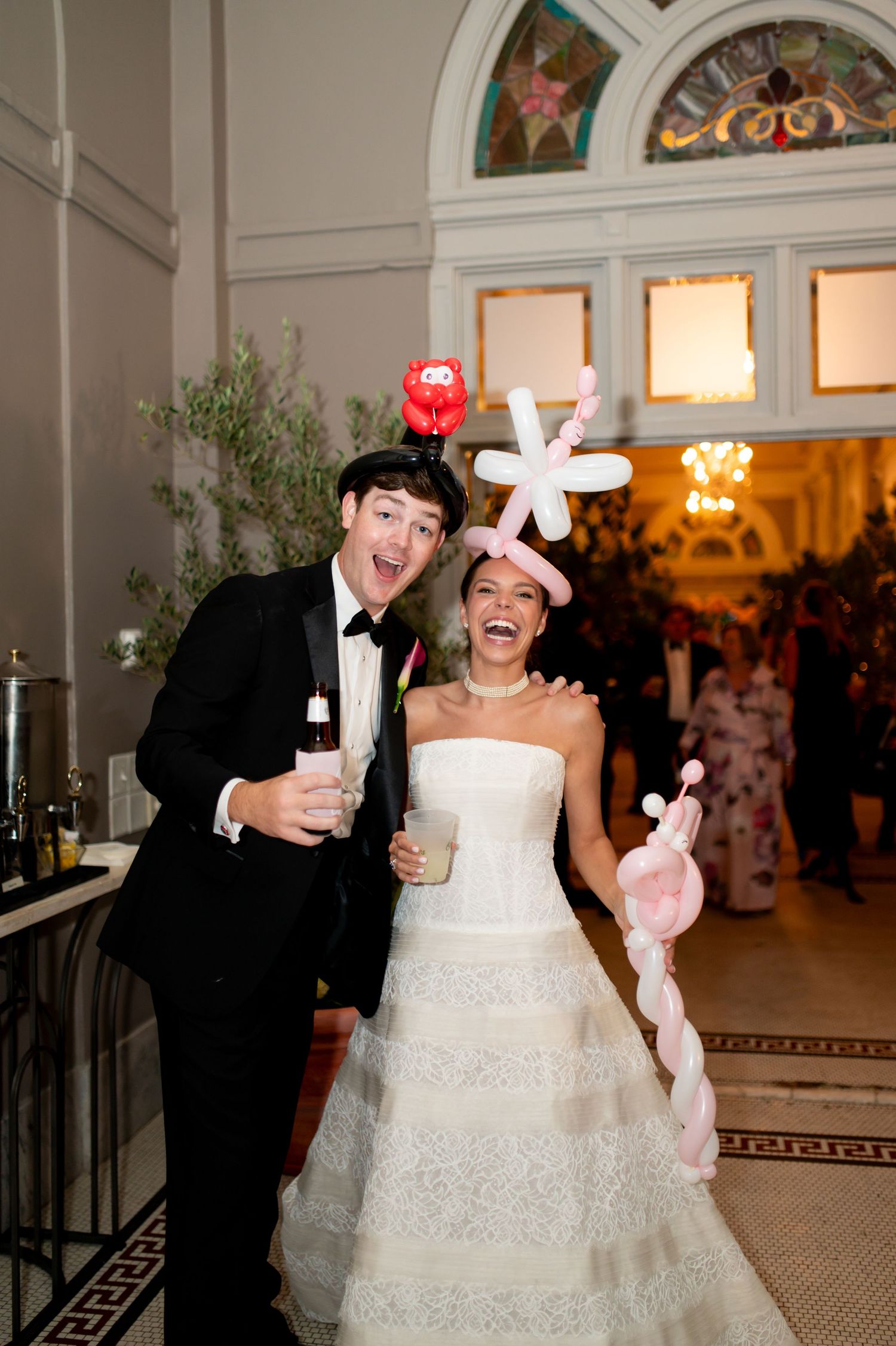 Joyful bride and groom wear balloon animal hats at indoor wedding reception with stained glass windows.