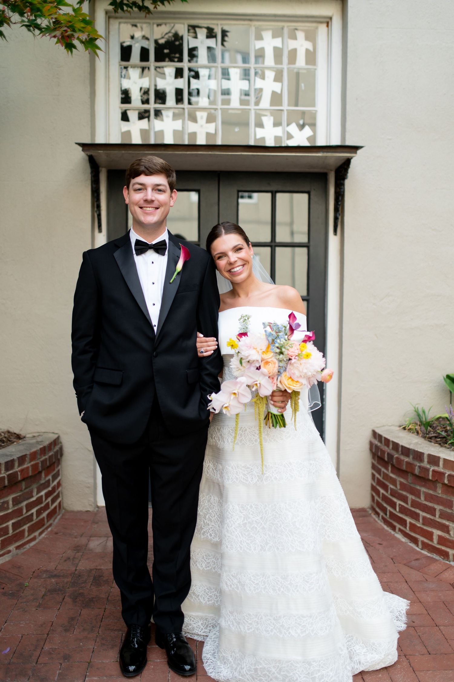 Bride in off-shoulder lace gown holding colorful bouquet stands with groom in black tuxedo.