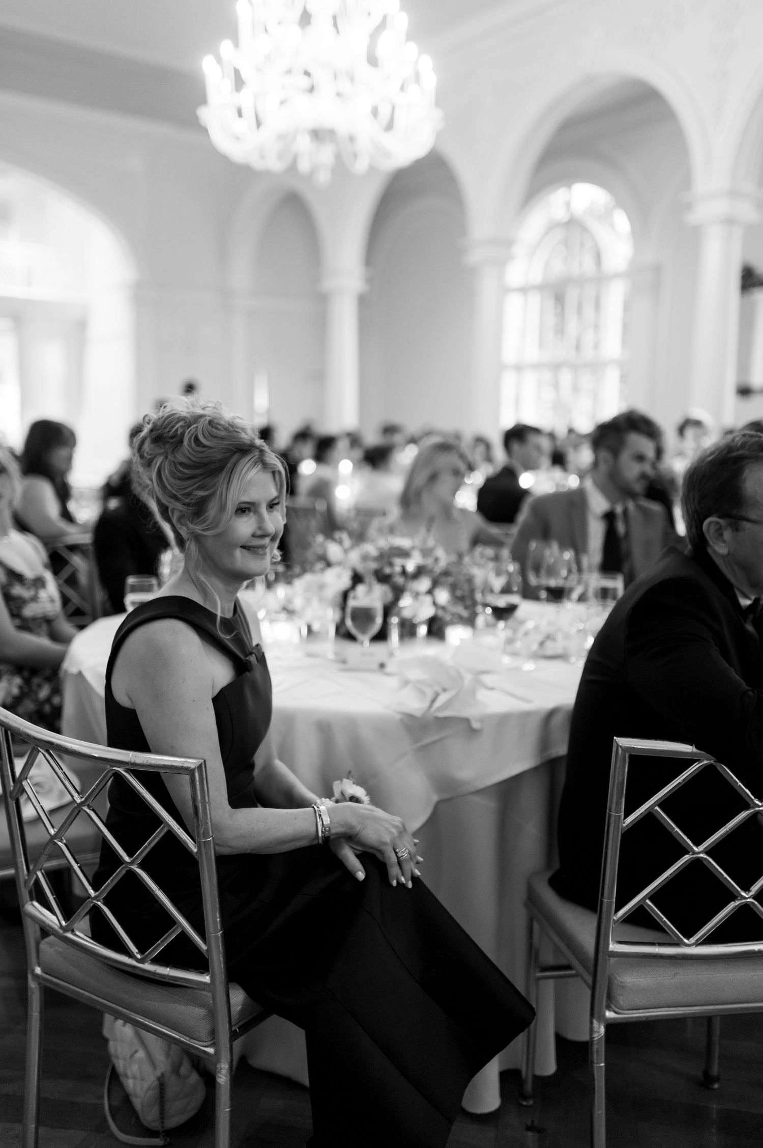 Wedding guests seated in elegant chairs at round tables beneath crystal chandeliers.