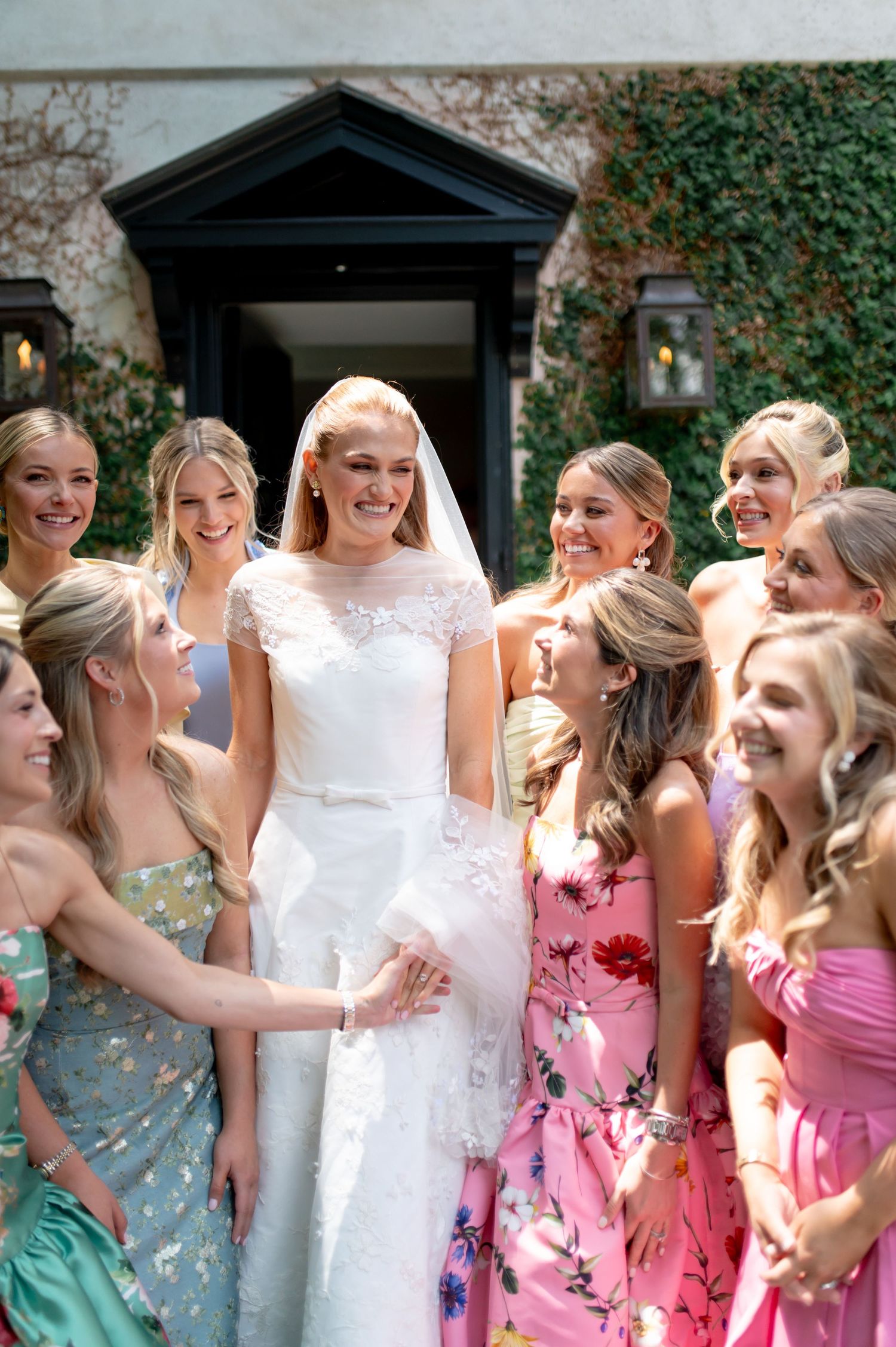 Smiling bride in lace cap-sleeve gown surrounded by bridesmaids in floral and pastel dresses outdoors.