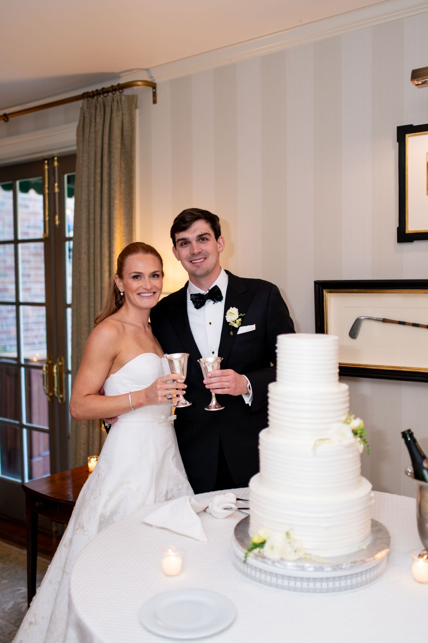 Bride and groom toast with champagne beside elegant four-tier white wedding cake at reception.