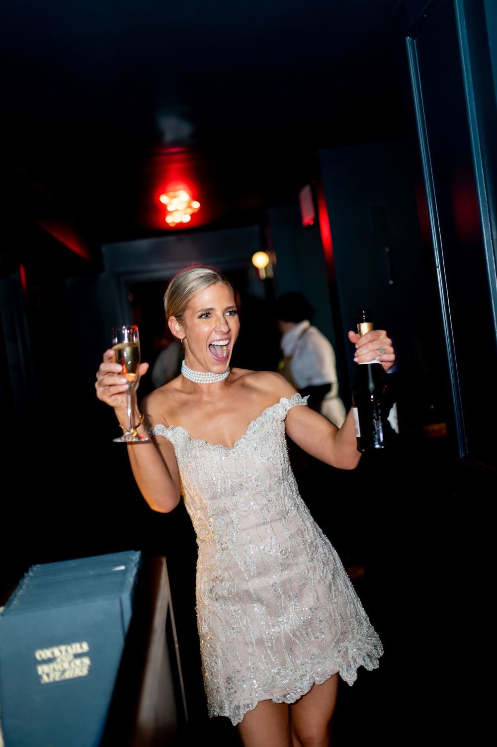 Excited bride in short beaded white dress holding champagne glass and bottle at wedding reception.