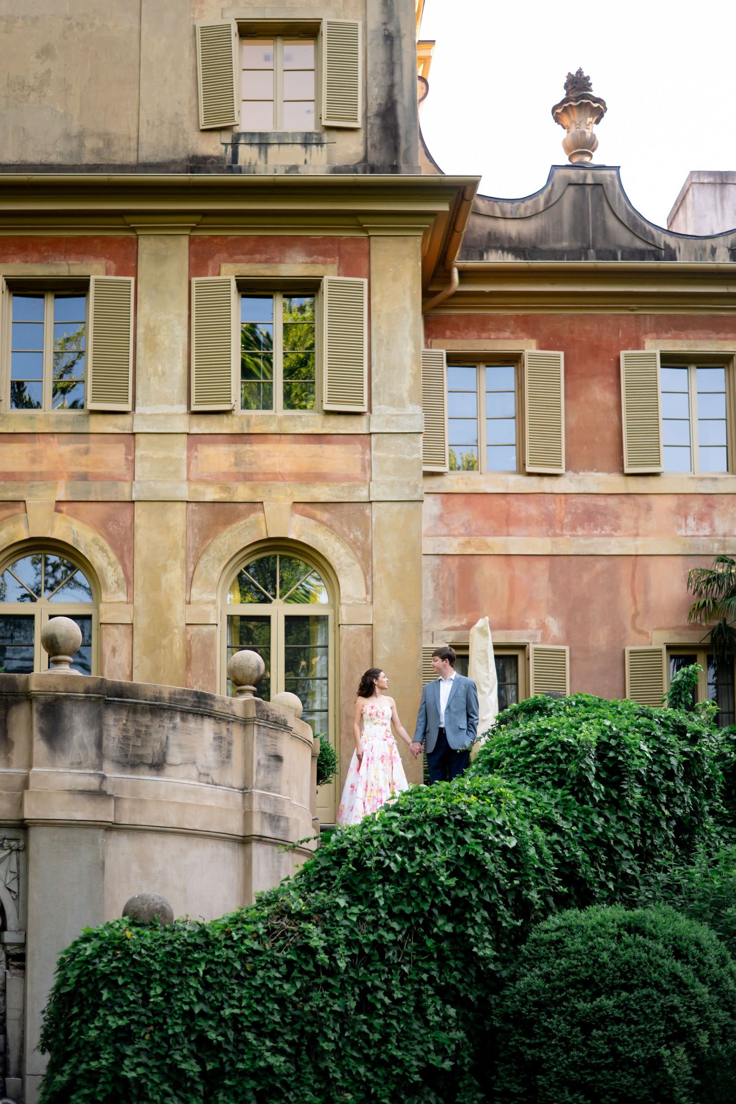 Couple in elegant attire standing on stone steps beside manicured hedges before a grand Italian villa.