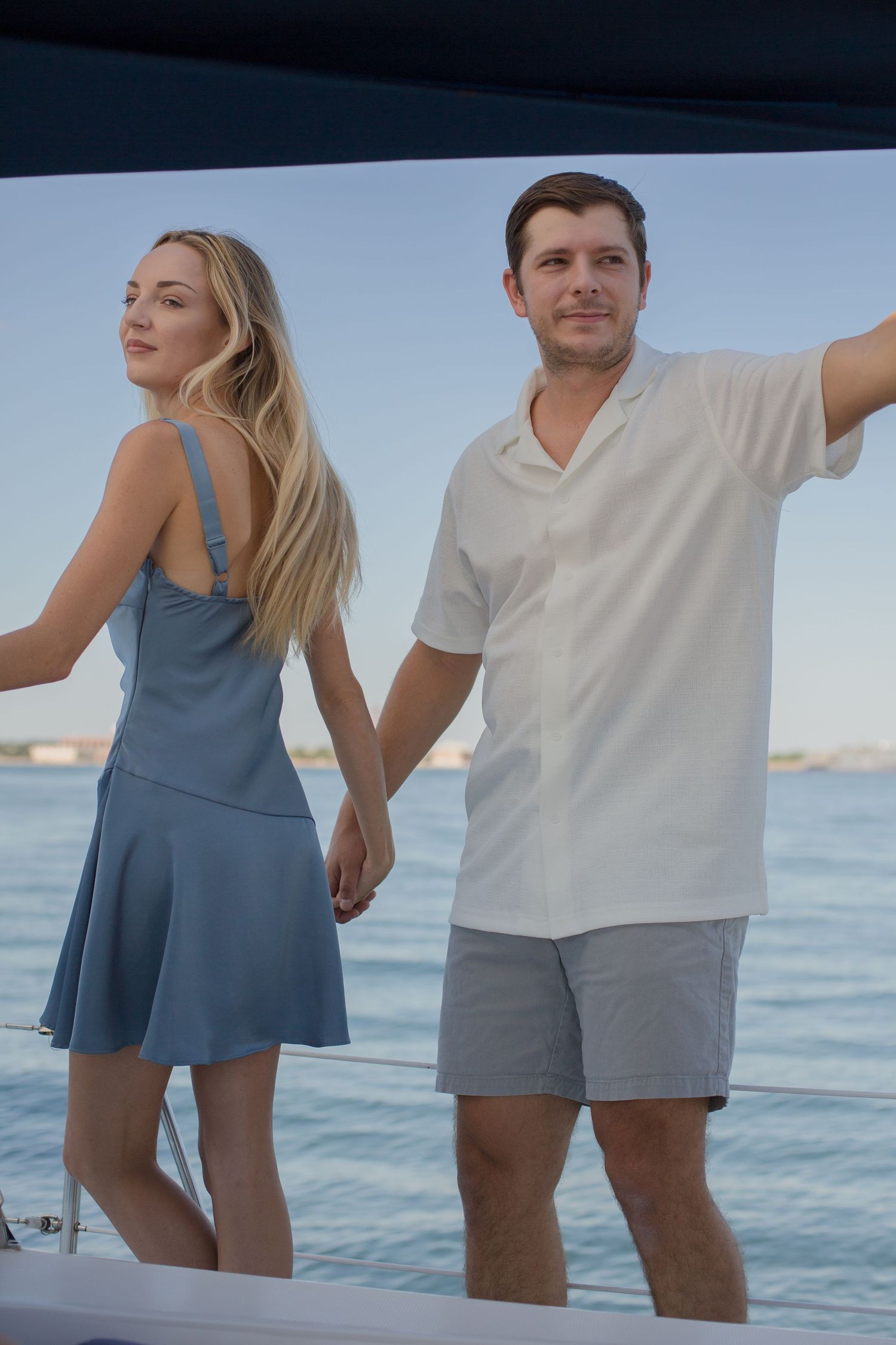 Couple holding hands standing on a sailboat deck with ocean views at dusk.