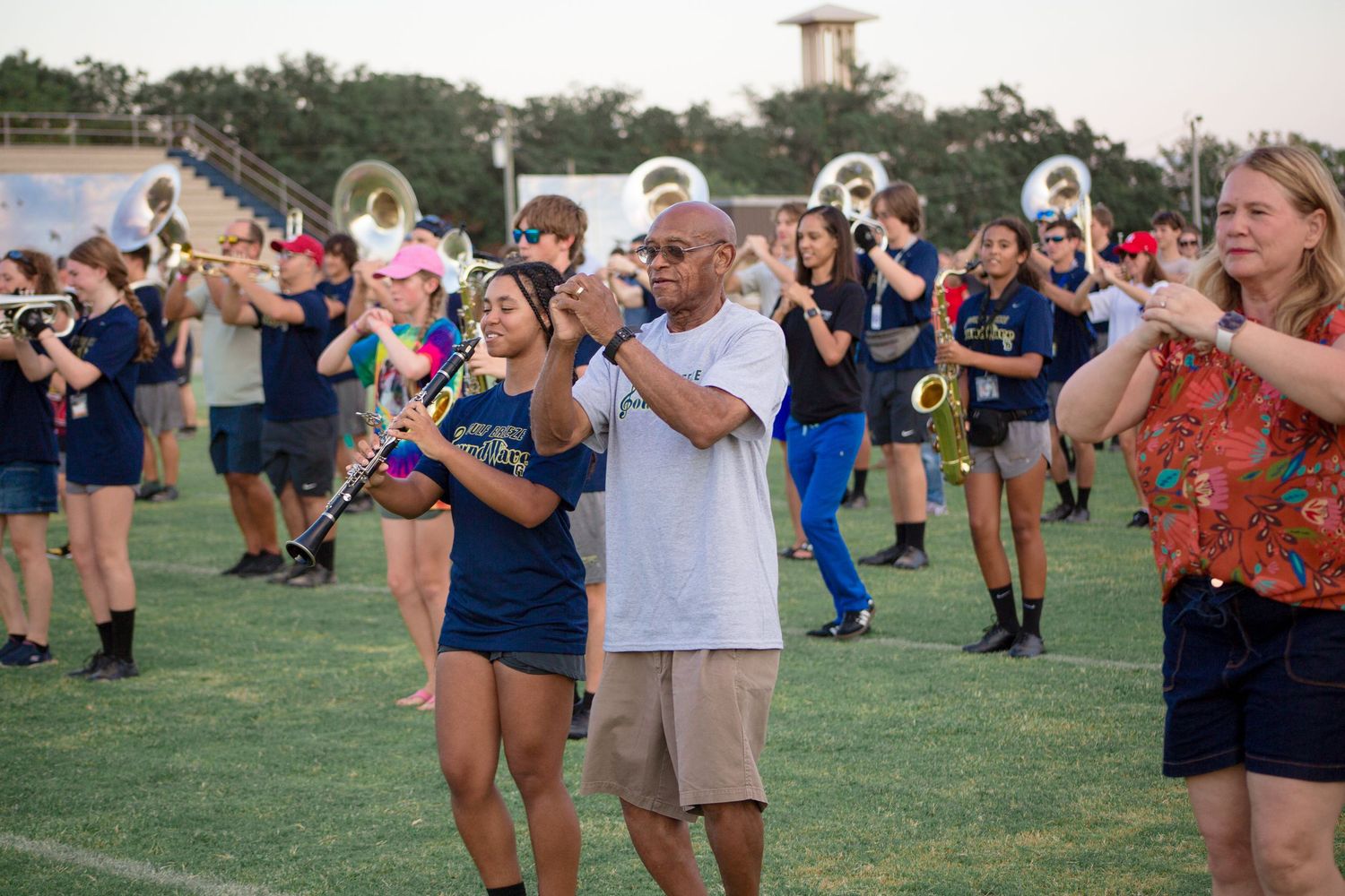 Marching band students practice on a field in navy uniforms, playing brass and woodwind instruments outdoors.