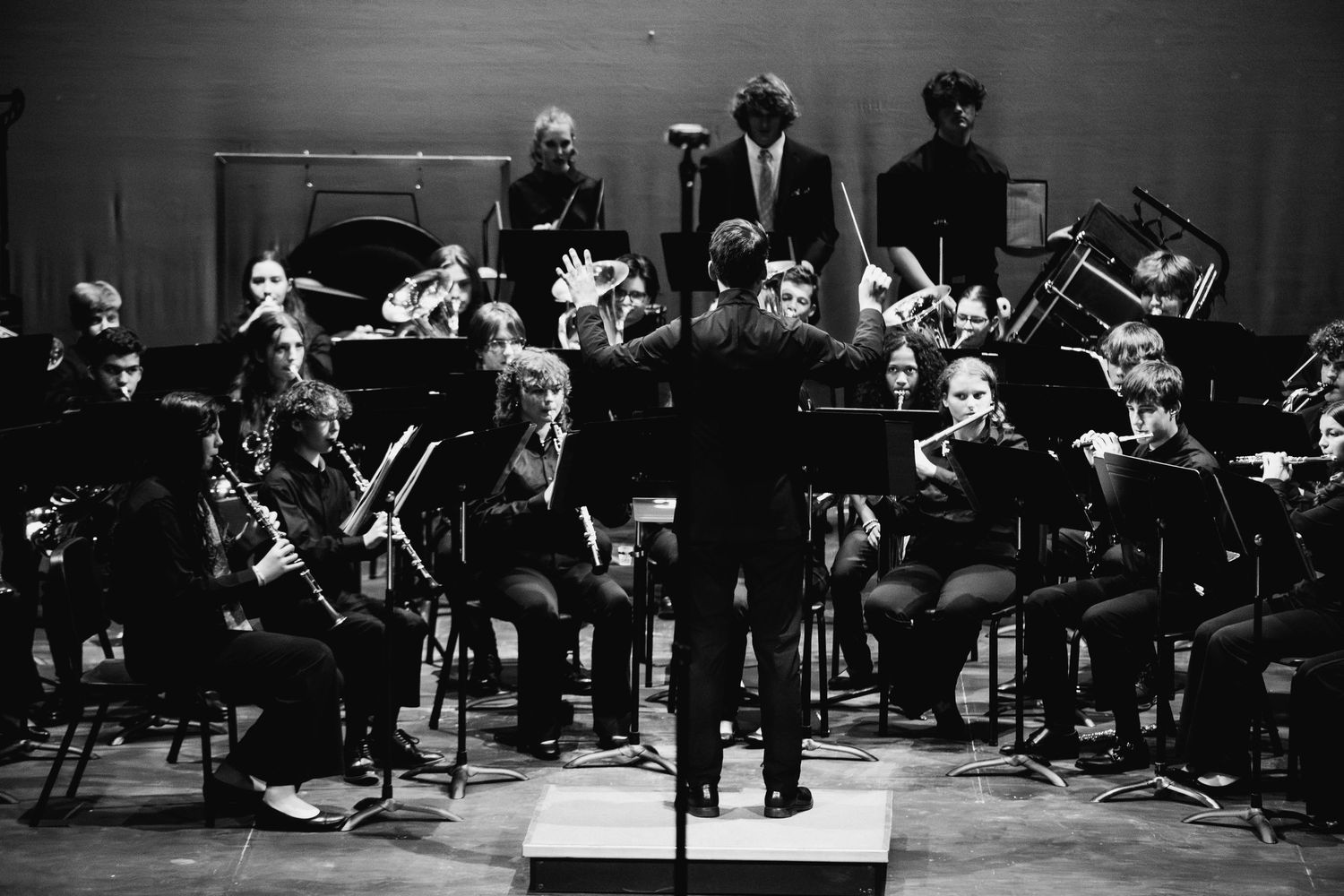 Black and white photo of a student orchestra performing on stage with a conductor leading the ensemble.