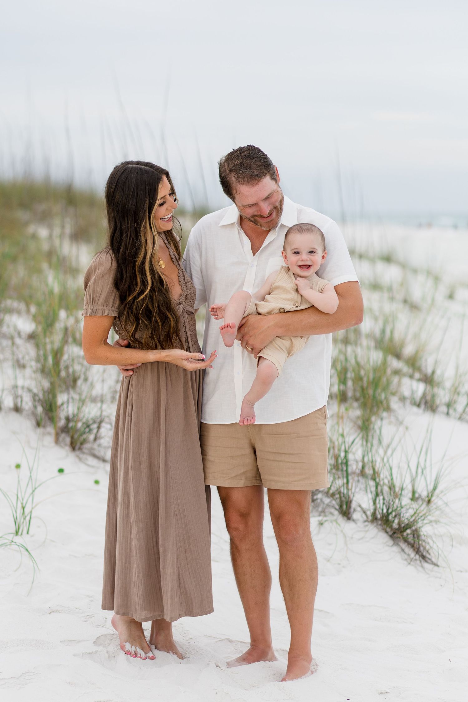 Happy young family with a smiling baby posing on white sand dunes at the beach.