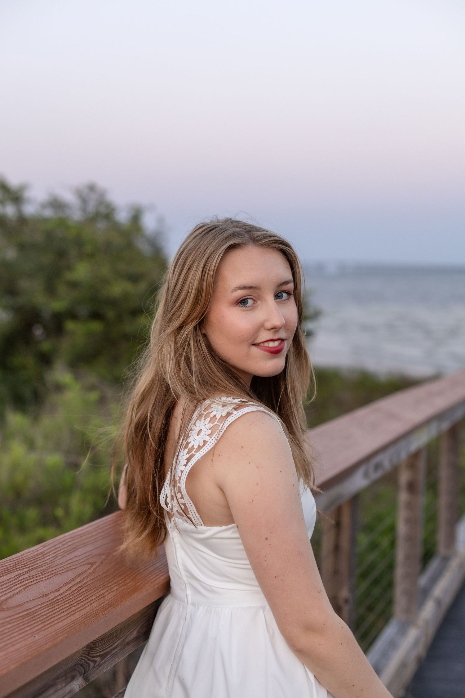 Elegant teen girl in a white dress posing on a beachside boardwalk at dusk.