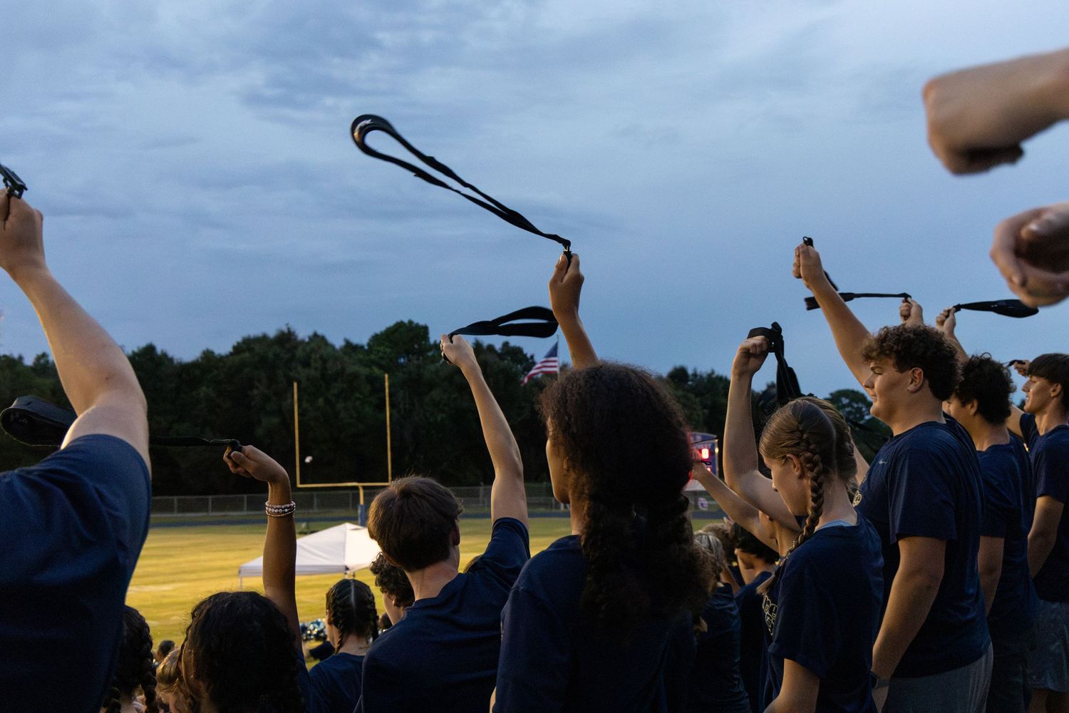 Students in navy shirts raise their hands holding black ribbons at a twilight outdoor event near a football field.