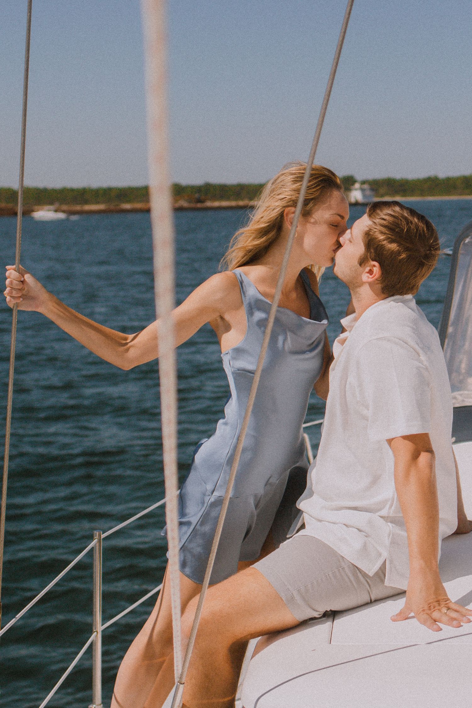 Couple kissing on a sailboat deck with blue ocean water and green shoreline in background.