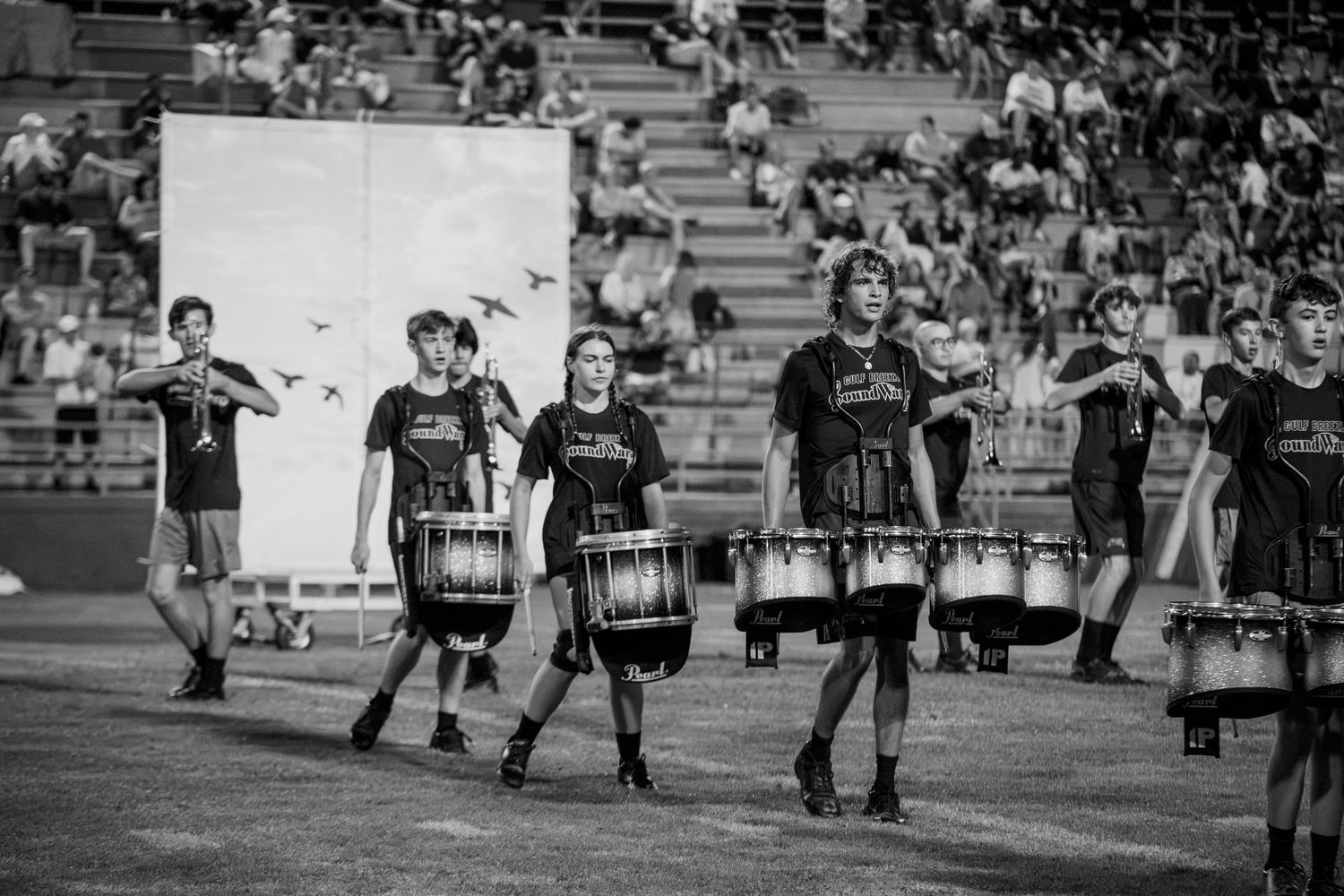 Black and white photo of marching band drummers performing on a football field during a halftime show.
