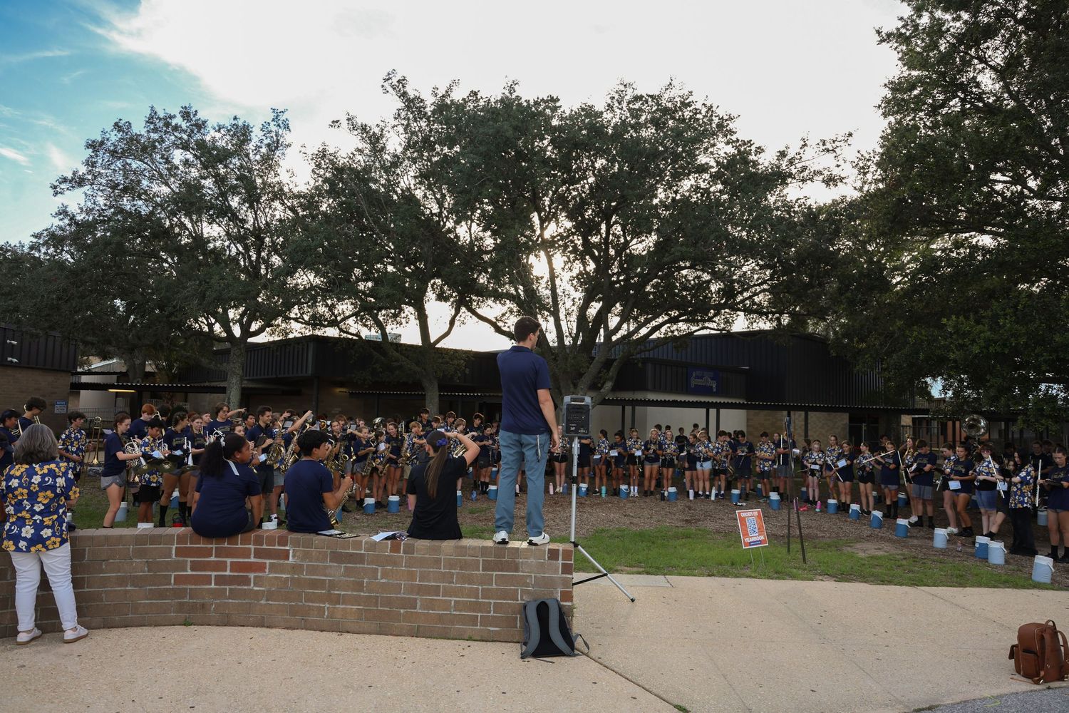 A speaker addresses a large crowd of students gathered outdoors at a school event on a sunny day.