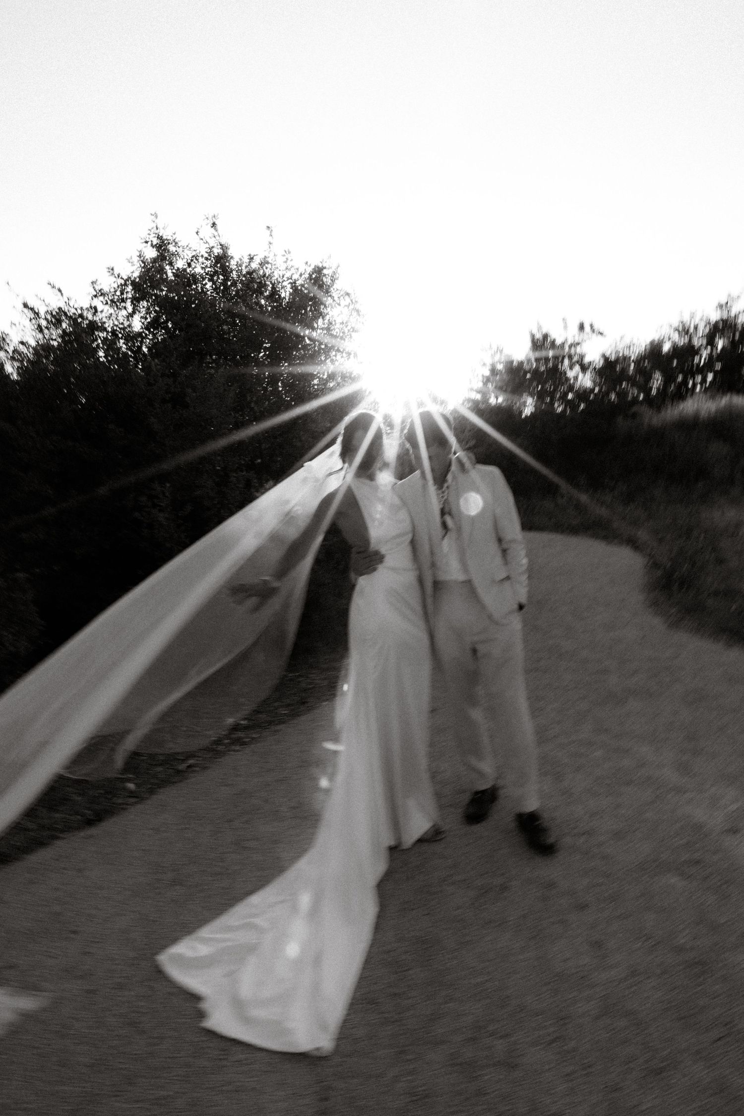 A couple in wedding attire poses dramatically in black and white with flowing veil against sunset.