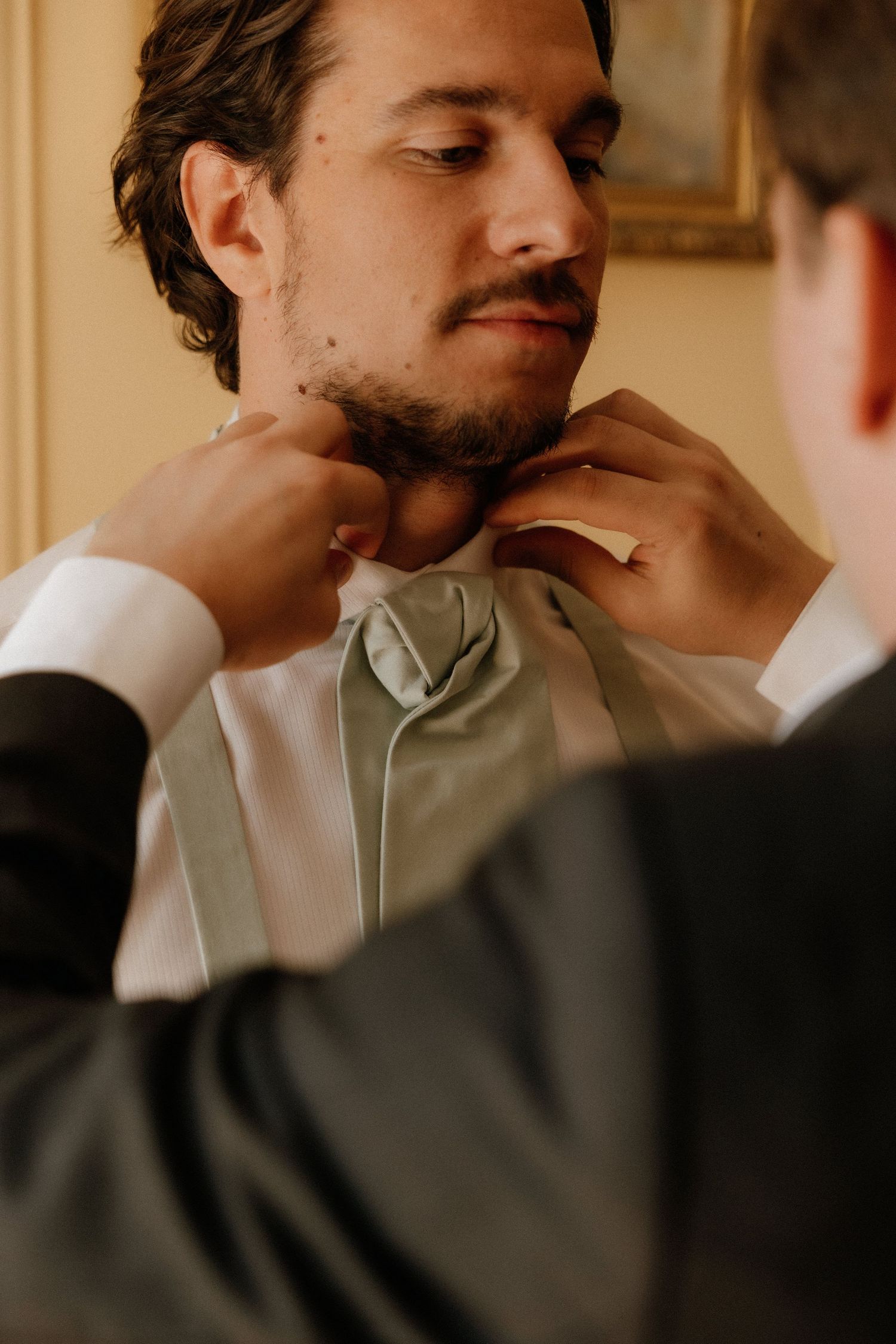 A bearded man in a light colored shirt sits thoughtfully with his hand near his face.