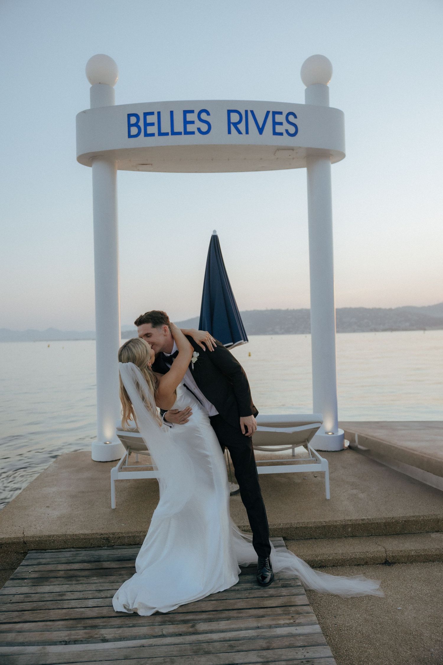 A couple shares a romantic wedding moment under a white archway labeled 'Belles Rives' on a waterfront dock at sunset.