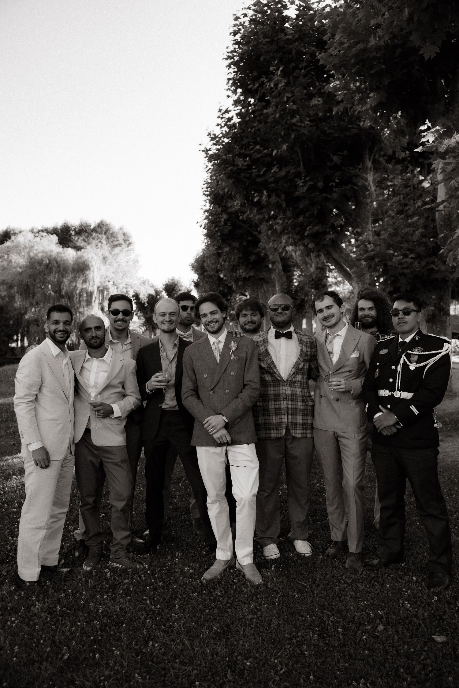 A black and white photo of a group of people in formal attire standing outdoors under trees.