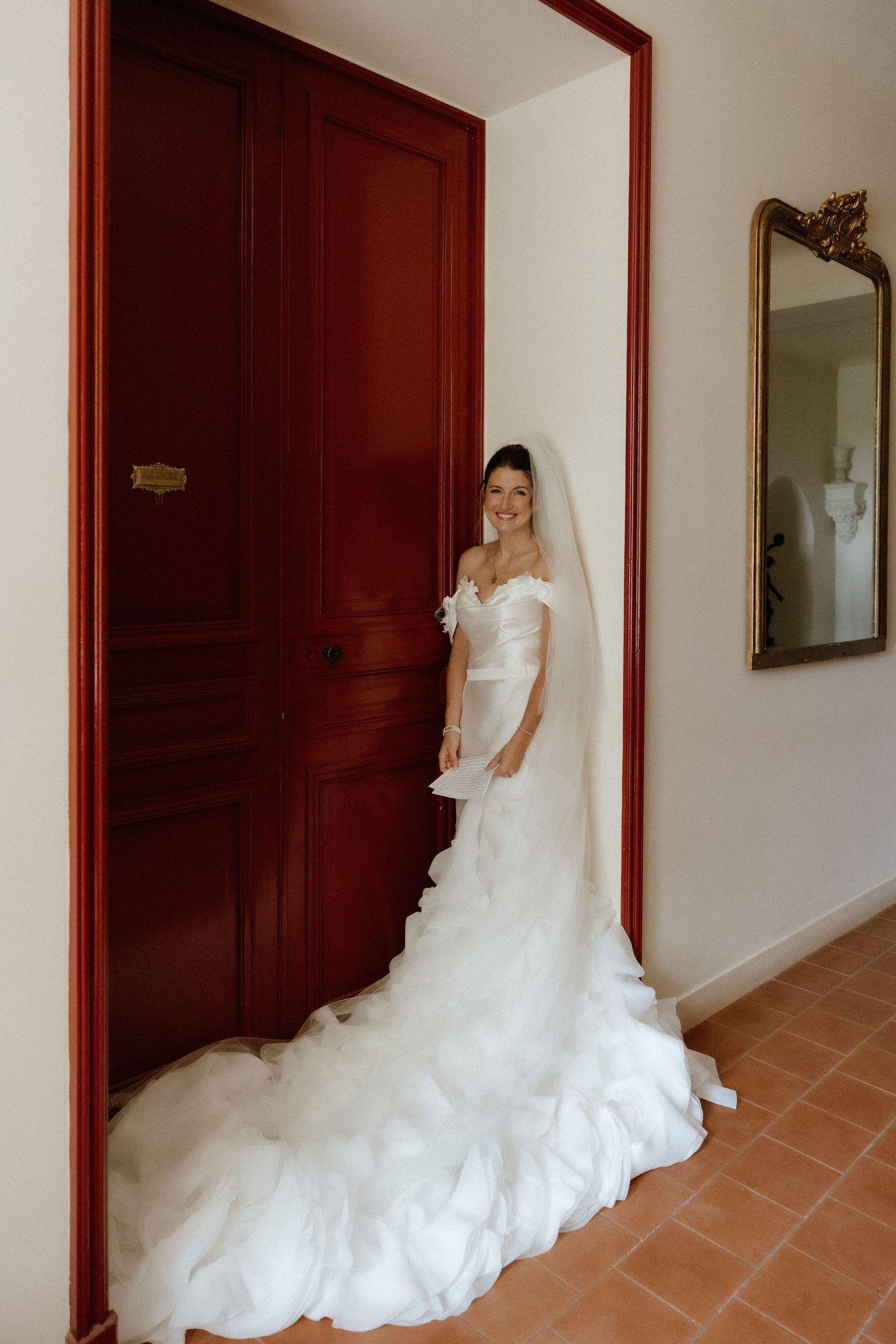 A bride in a flowing white wedding gown poses elegantly against dark wooden doors in a hallway.