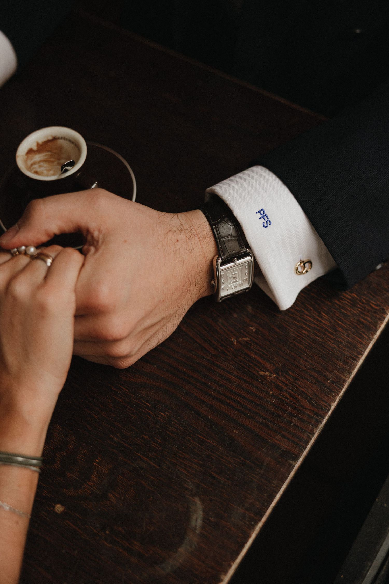 Close-up view of hands wearing monogrammed white dress shirt cuffs on a dark wooden table.
