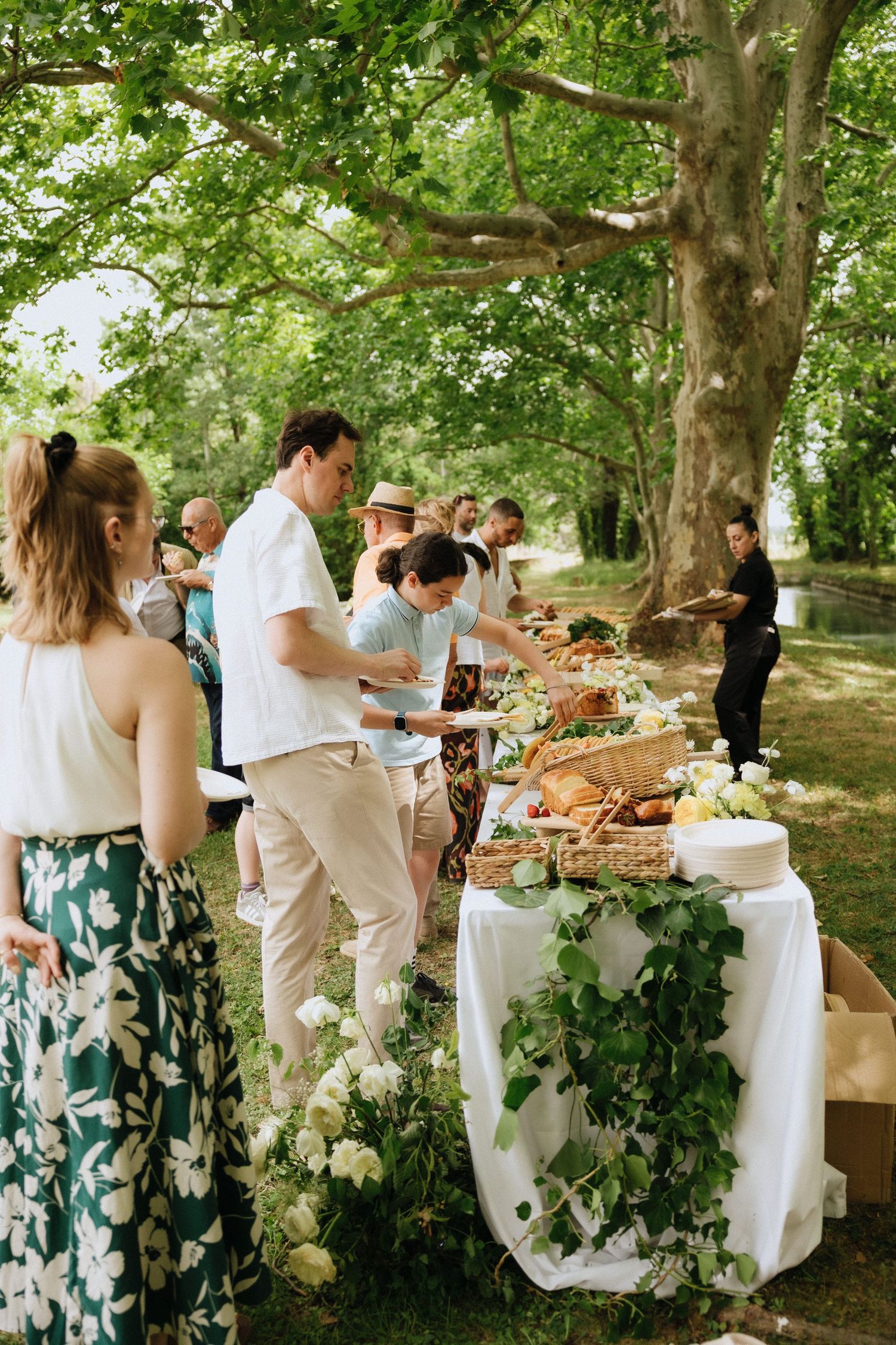 A buffet table decorated with greenery and white linens set up under trees for an outdoor wedding reception.