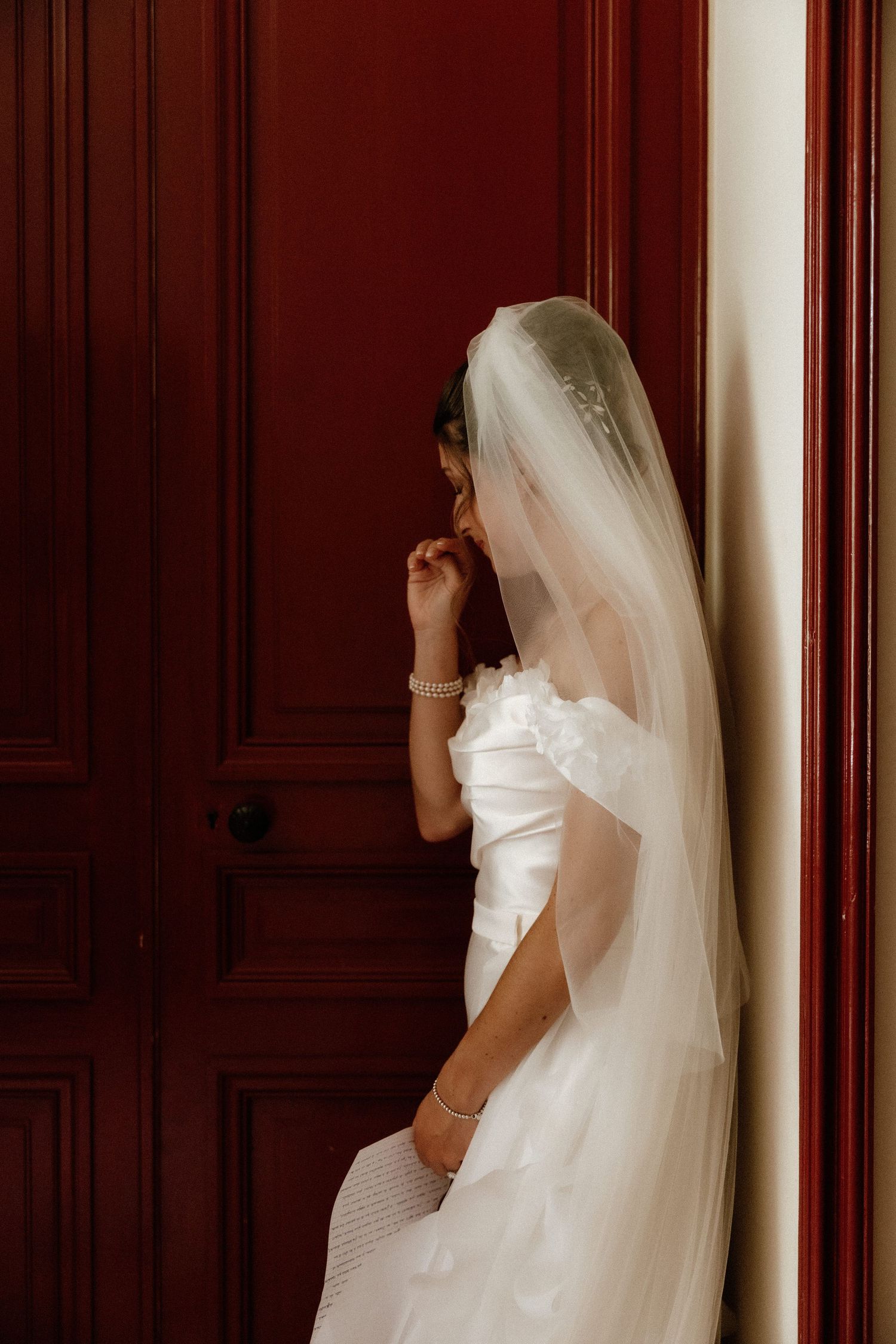 Bride in white dress and veil stands near wooden paneled wall.