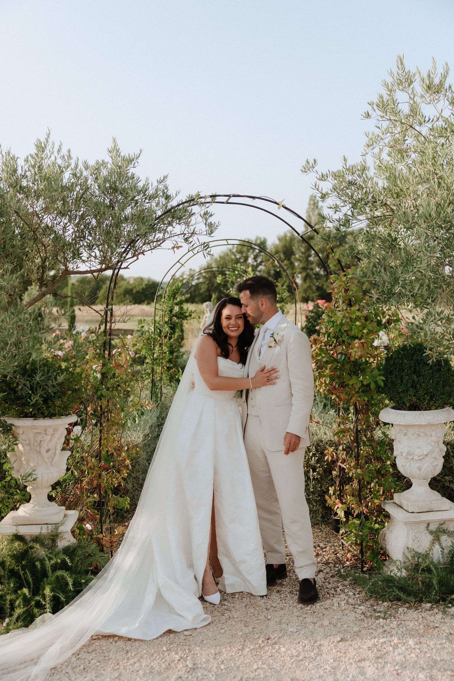 A romantic wedding couple shares intimate moments in a garden setting with decorative urns and olive trees.