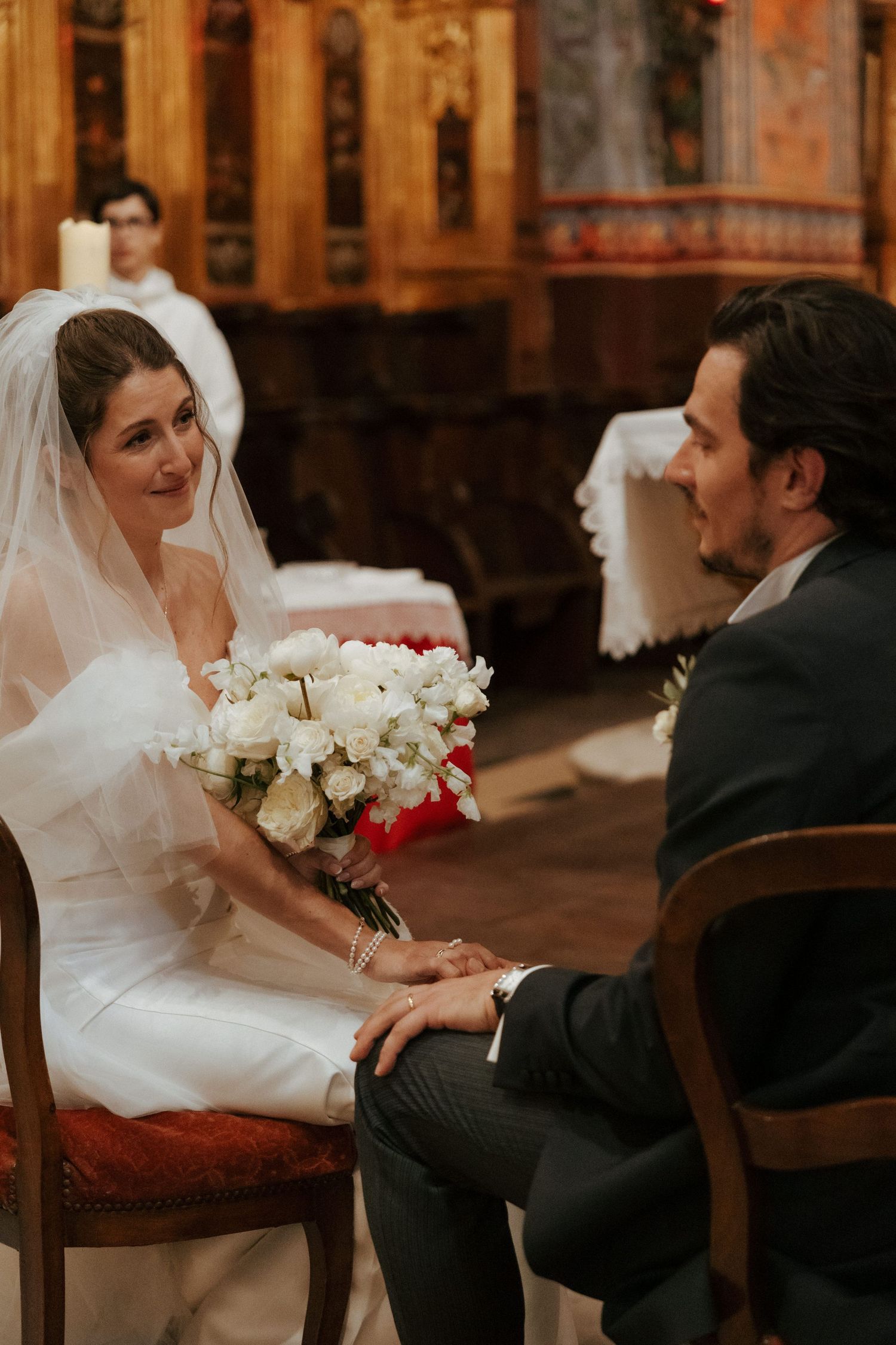 Wedding scene with couple seated indoors, featuring white floral bouquet and formal attire.