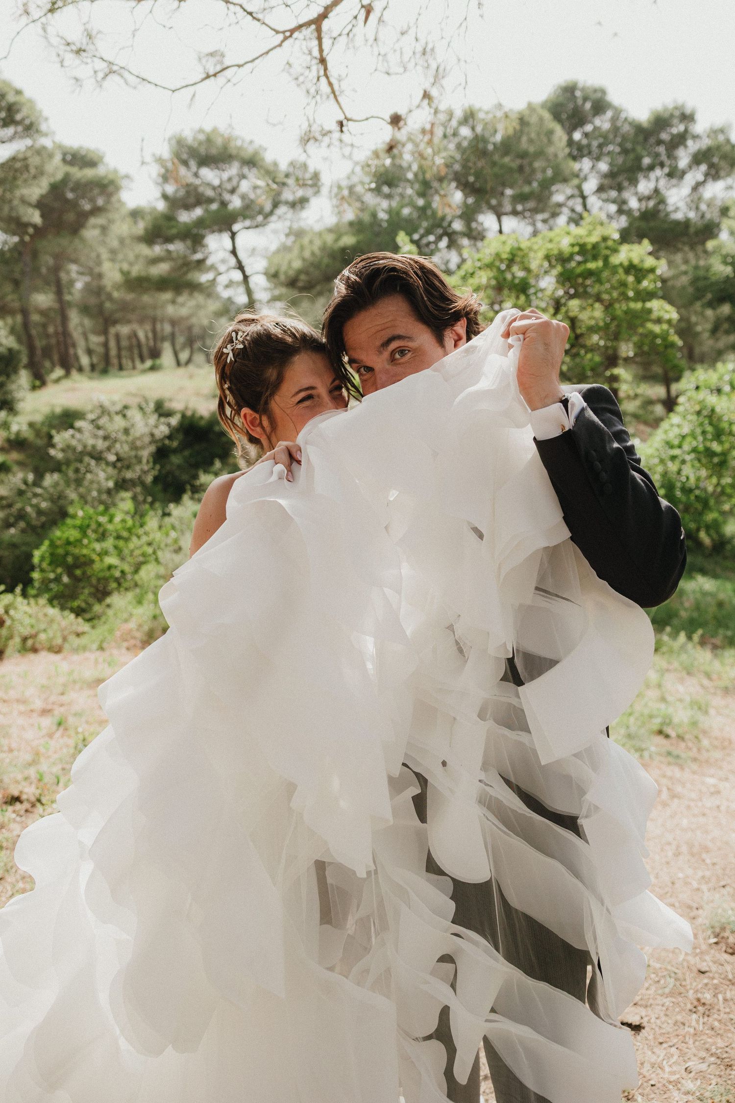 Playful wedding couple peek through flowing white wedding dress fabric in a sunny outdoor setting.