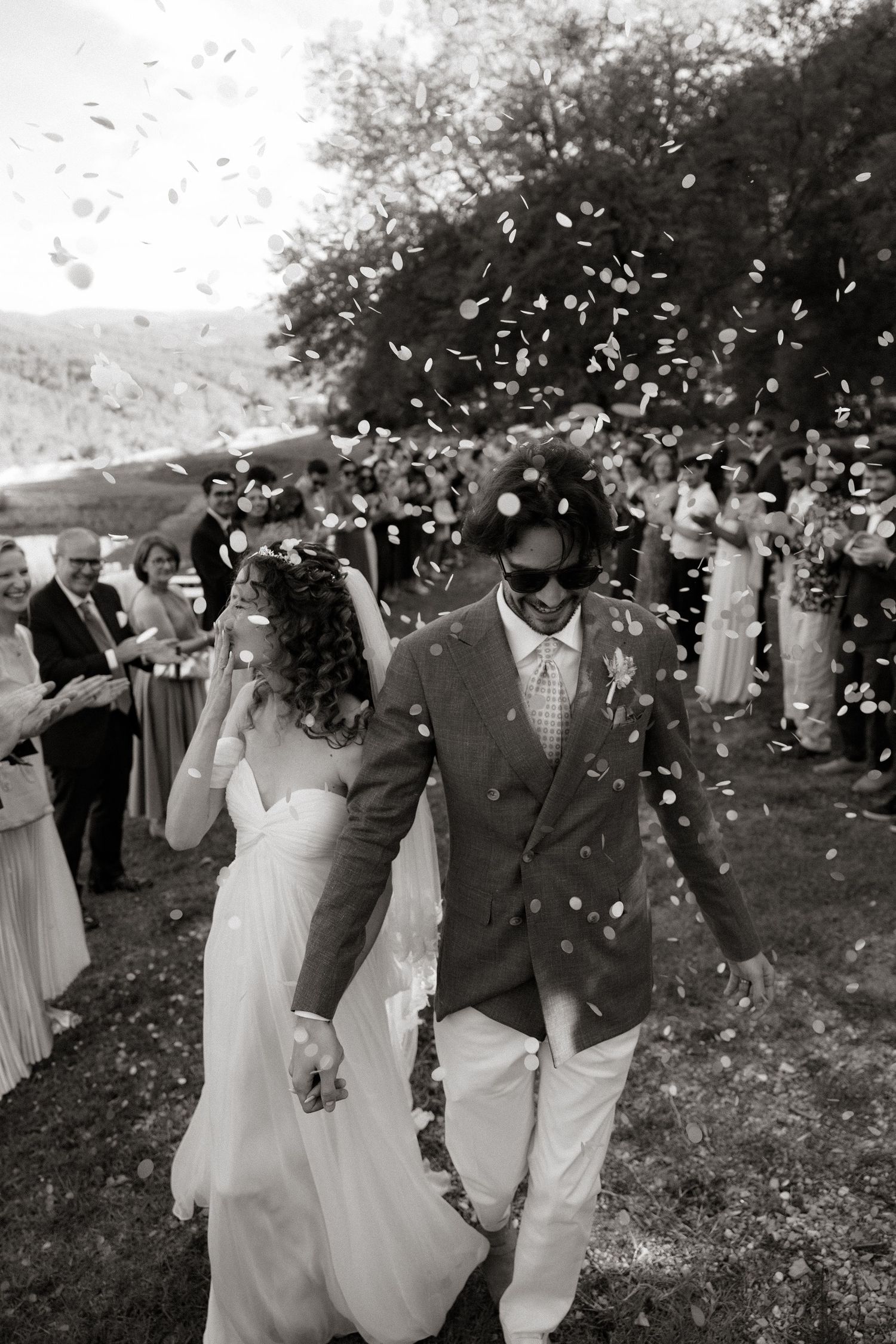 A couple walks through a shower of white confetti at an outdoor wedding ceremony while guests look on in celebration.