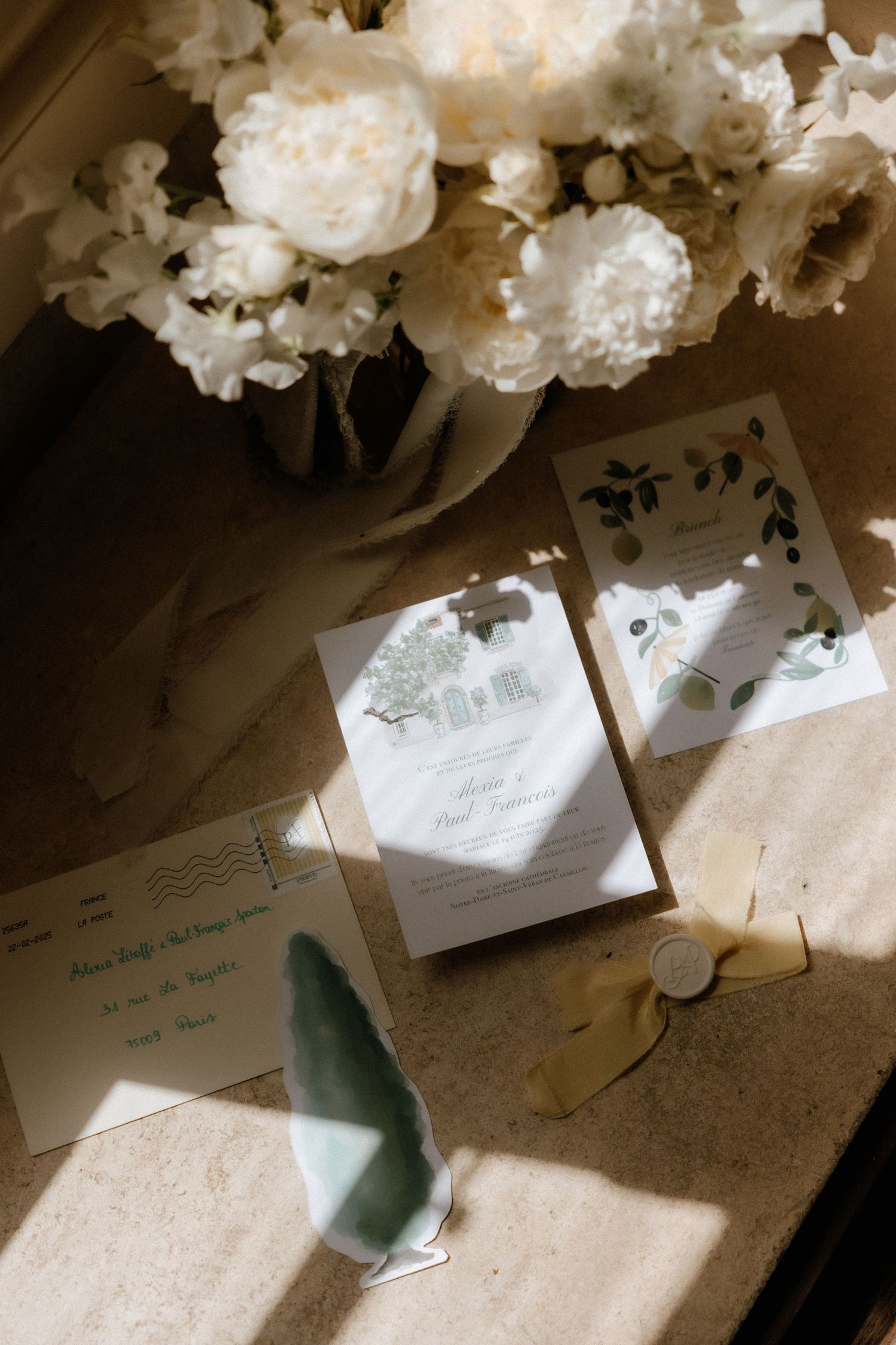 White flowers cast shadows over scattered wedding invitation cards on a light surface in natural sunlight.