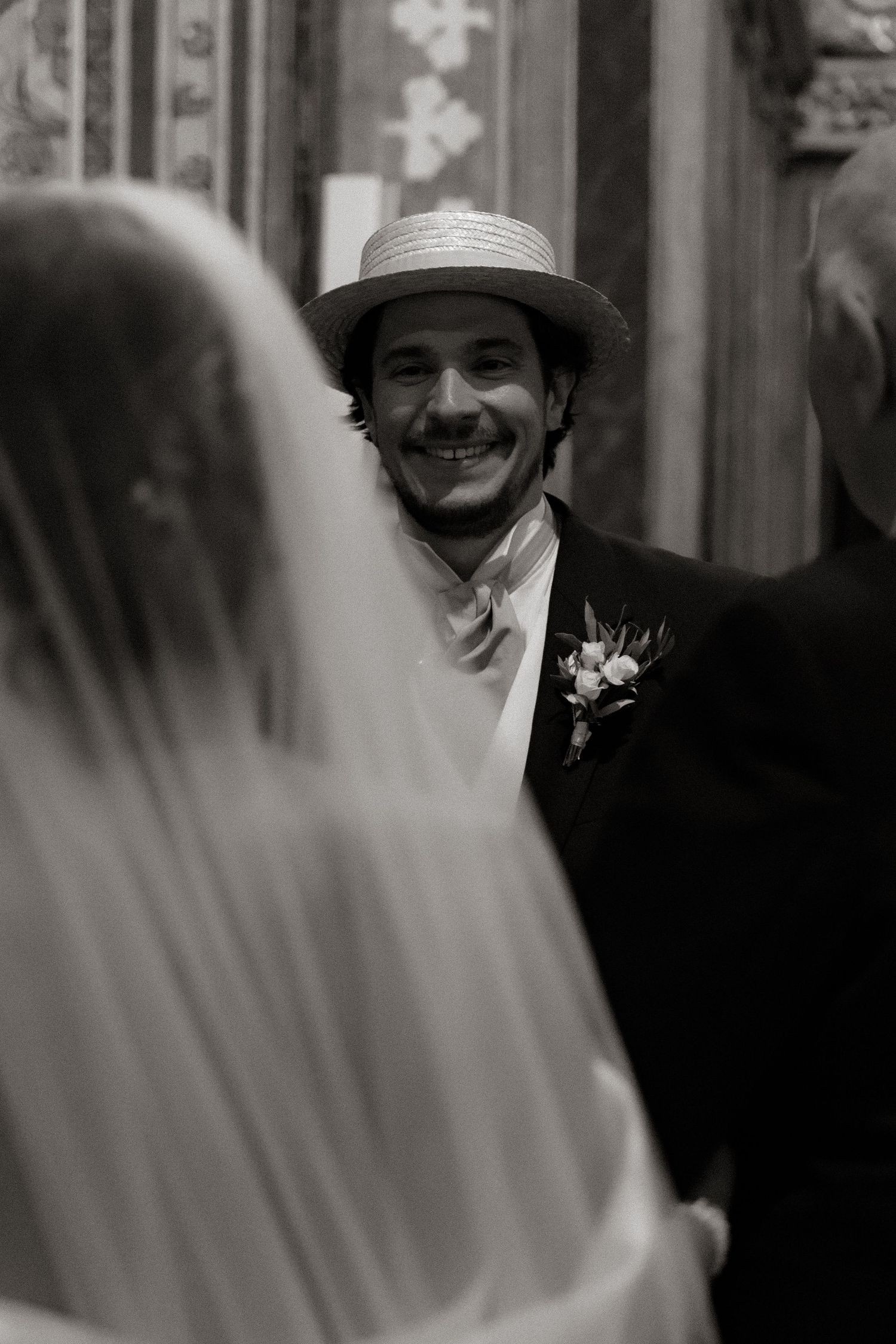 A smiling groom in a straw hat and formal attire stands with a bride in a veil.