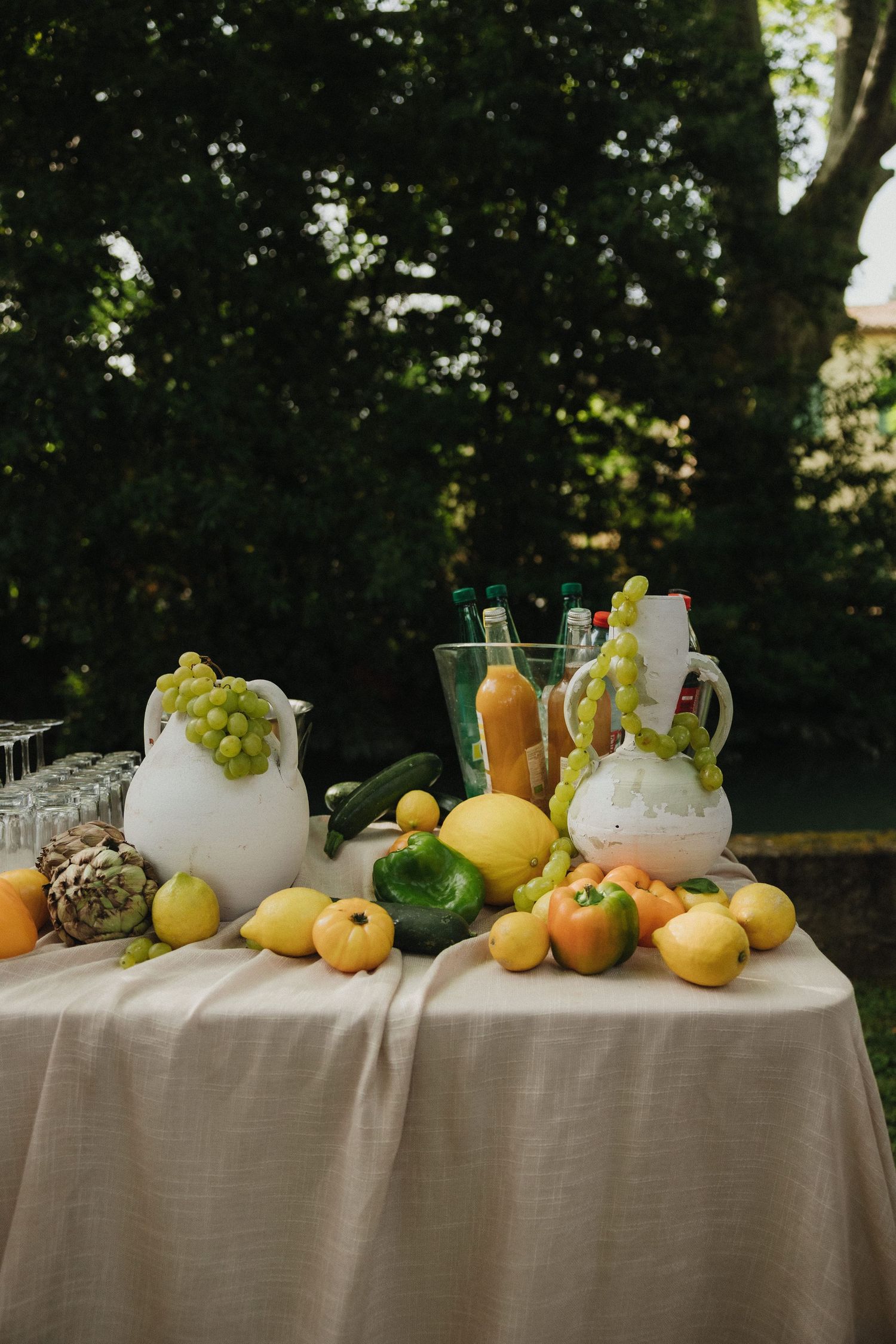 A rustic outdoor table display with fresh fruits, lemons, and glass pitchers arranged on a white tablecloth.