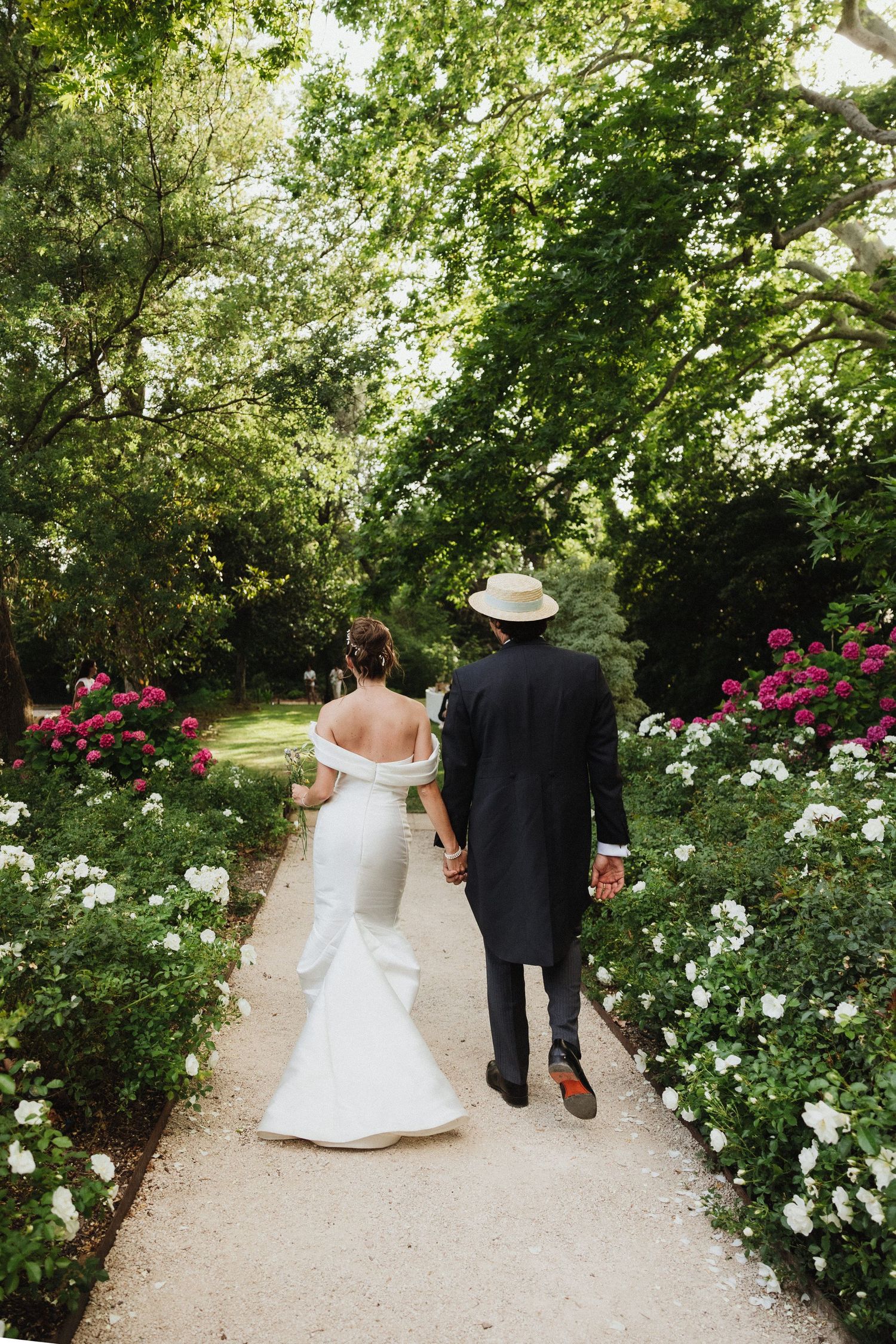 Newlyweds walking hand in hand down a garden path lined with blooming roses.