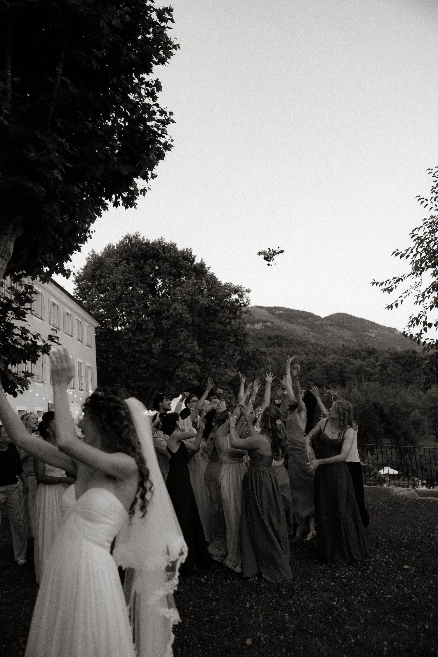 A group of wedding guests reach upward to catch the bouquet toss against a backdrop of mountains and trees.