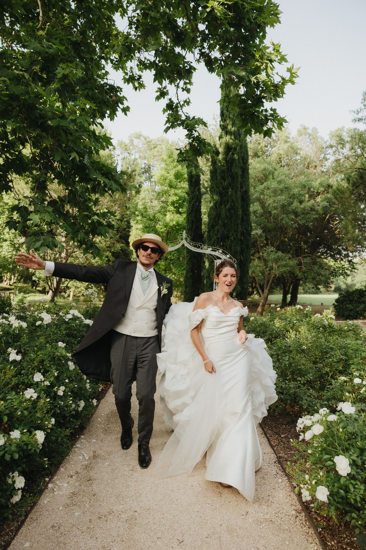 Newlyweds playfully walk and dance together on a tree-lined garden path.
