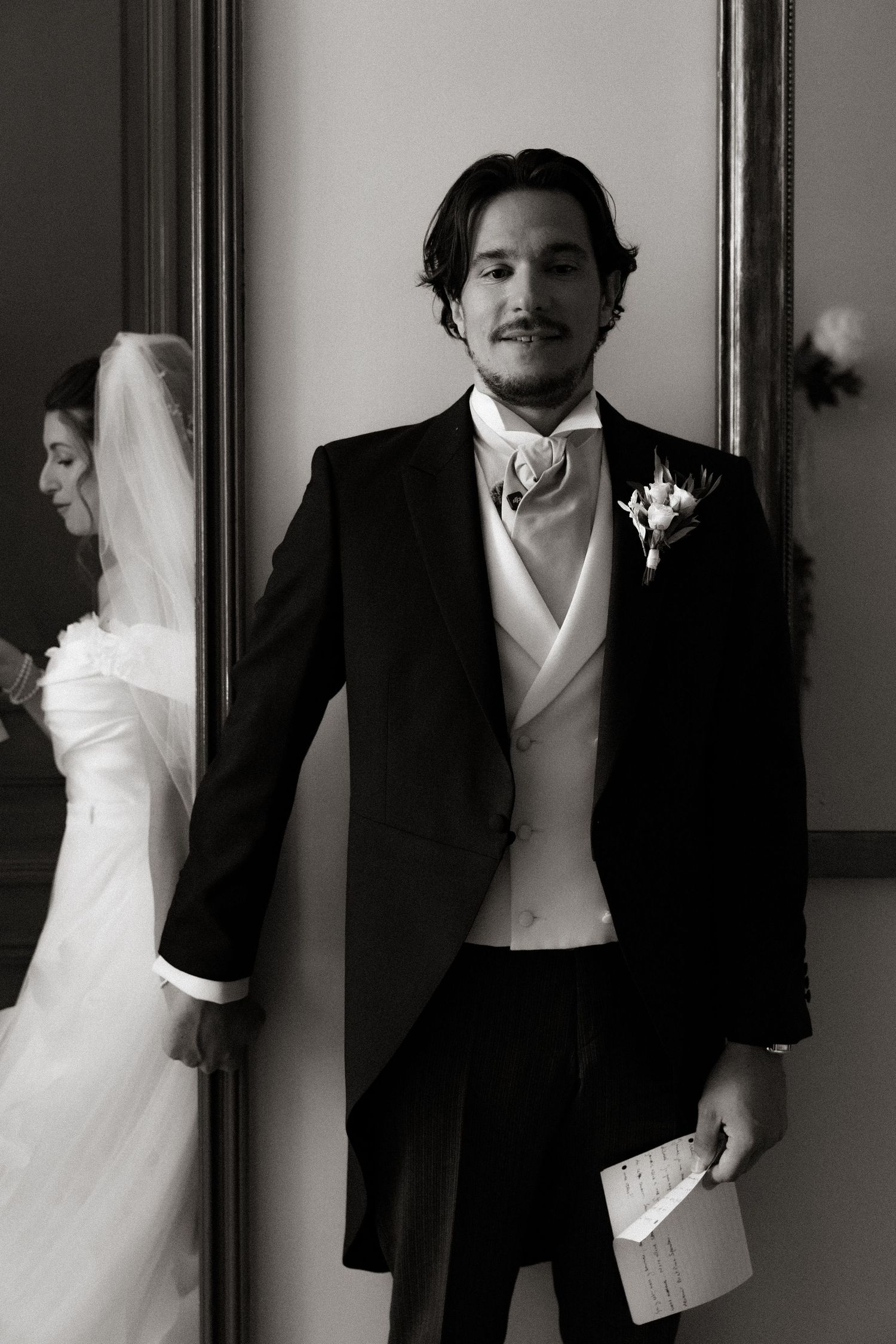 A groom in a formal black tuxedo and white vest poses elegantly while holding papers at a wedding.