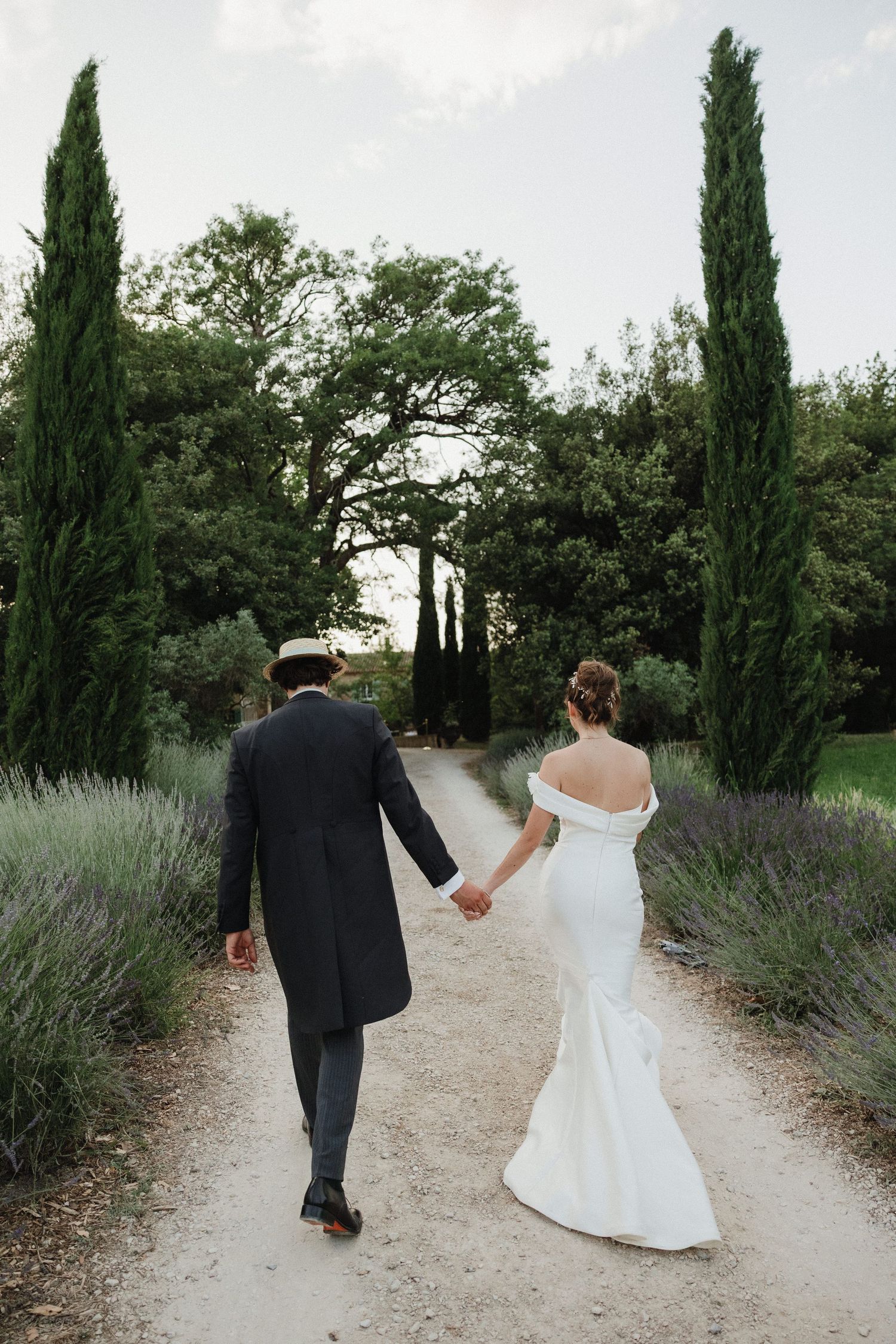 Elegant couple walking down a cypress-lined path with lavender bushes in a romantic garden setting.